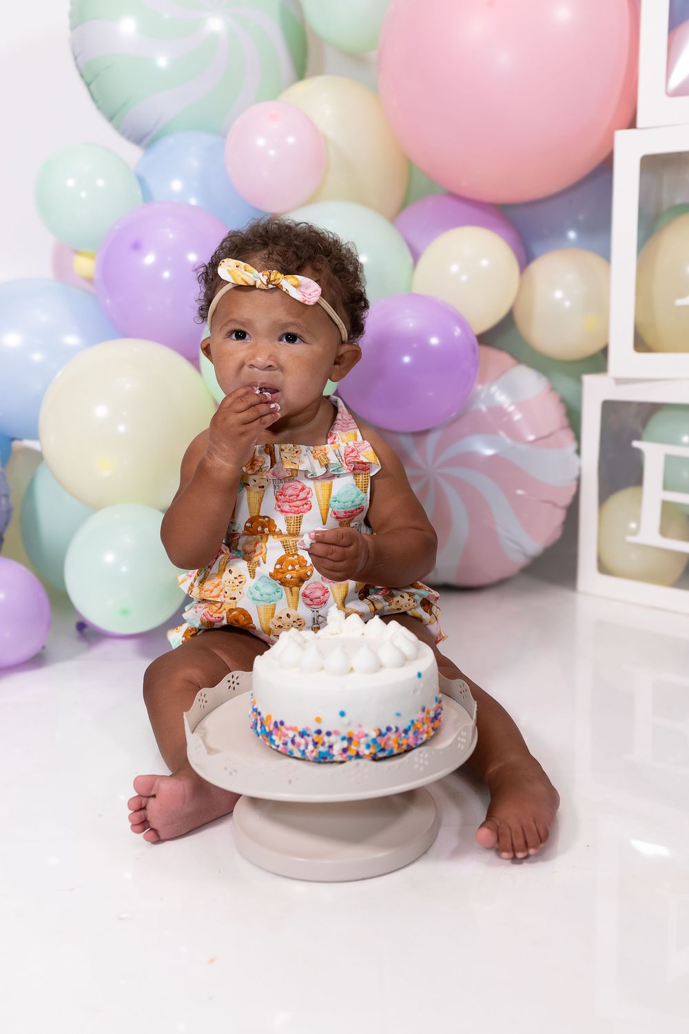 A baby in a colorful outfit sits next to a birthday cake with balloons in pastel colors decorating the background.