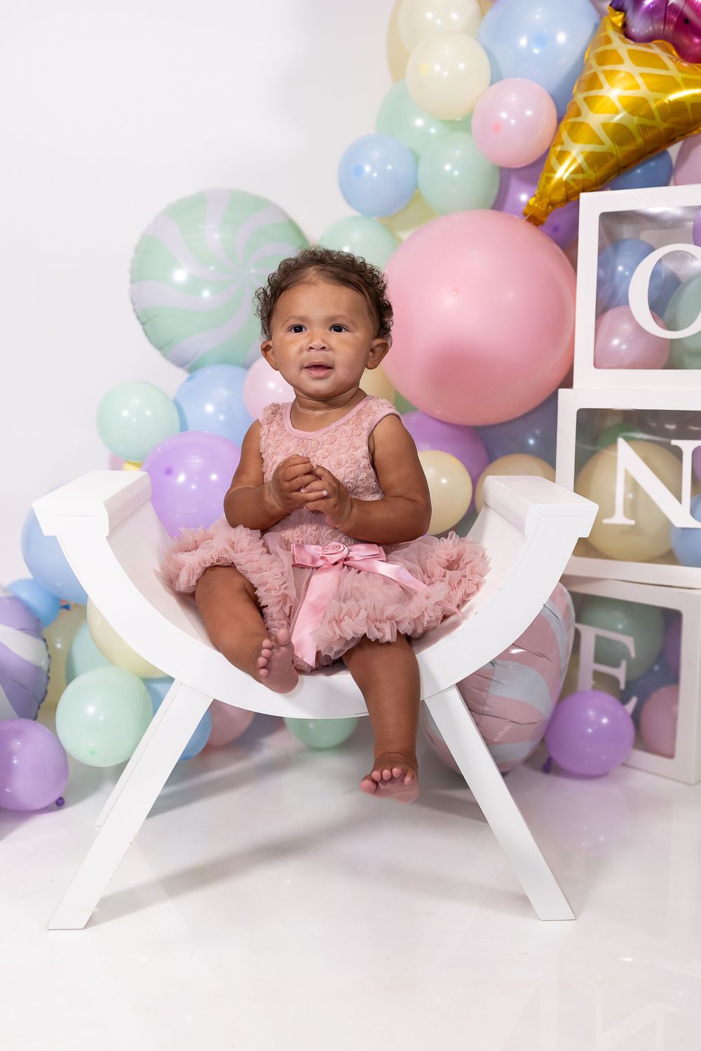 A small child in a pink tutu dress poses among pastel balloons and decorative letters during a photo session.