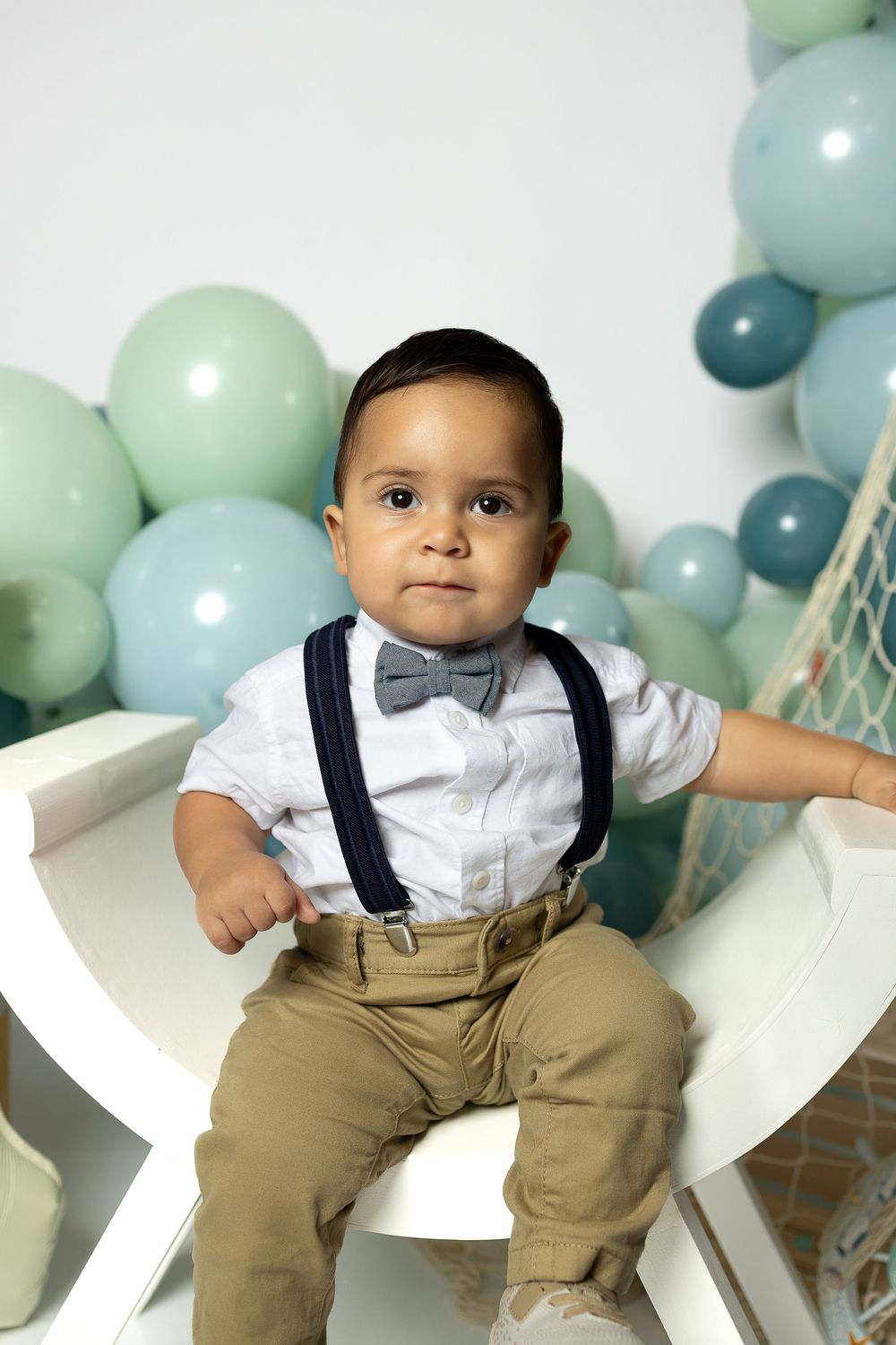 Baby in white dress shirt and khaki suspenders poses against blue and mint balloon backdrop for milestone photos.