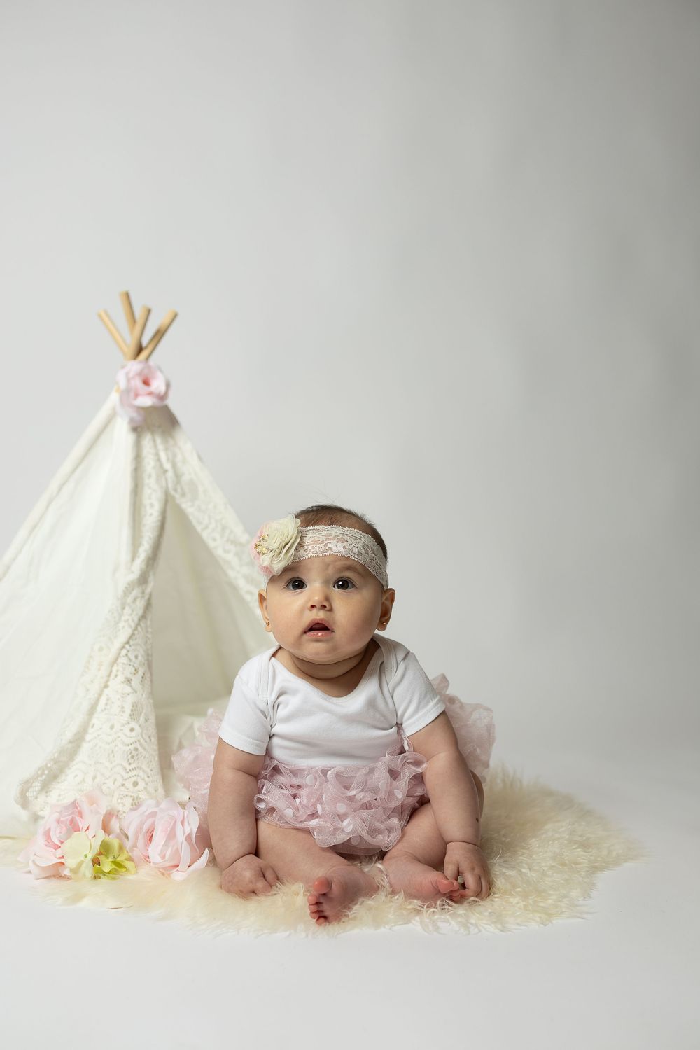 Baby in white outfit with pink ruffles sits between two decorative teepee tents against neutral background.