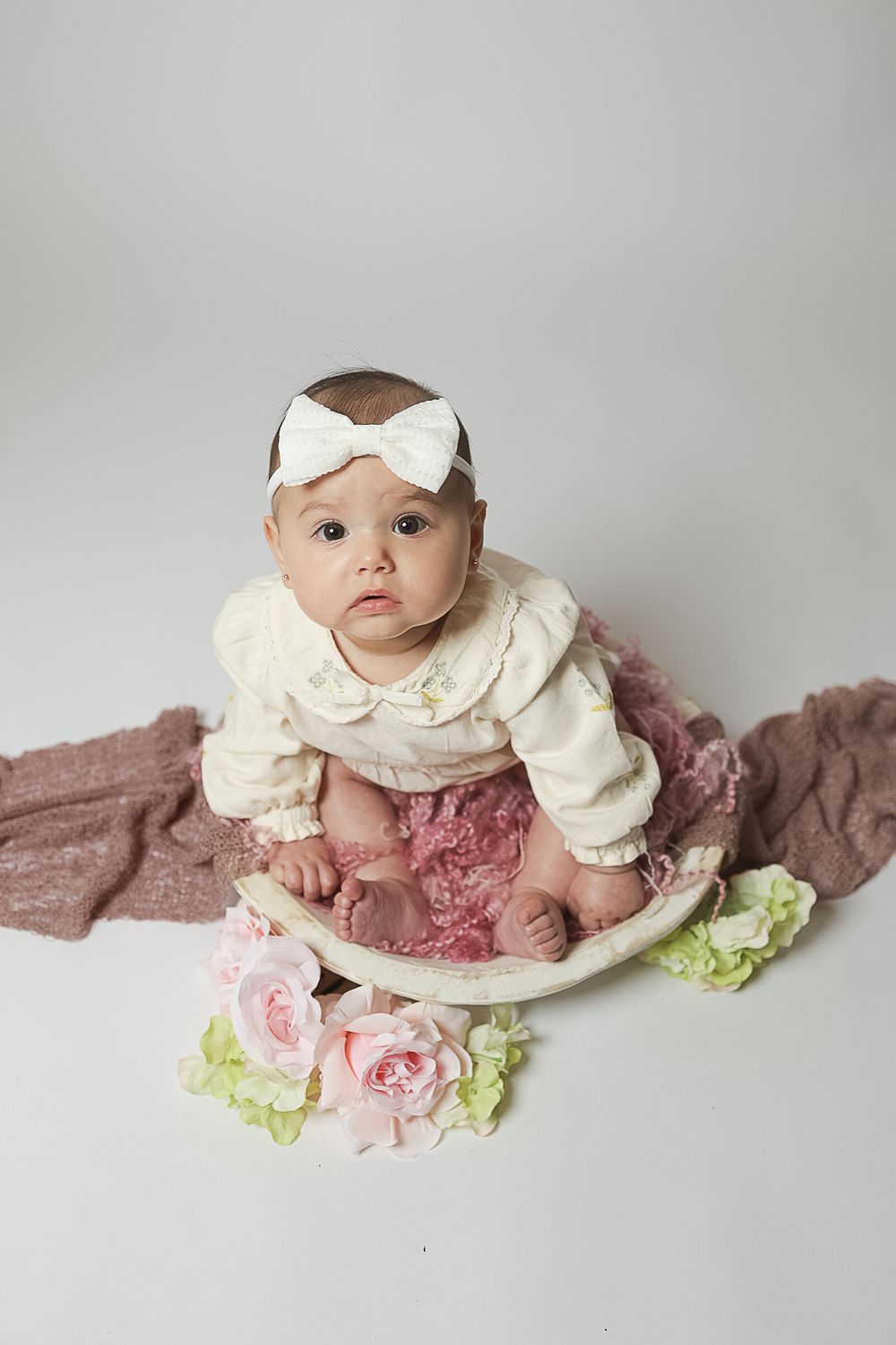 A baby in a cream-colored dress lies on a mauve blanket with pink and green floral decorations.