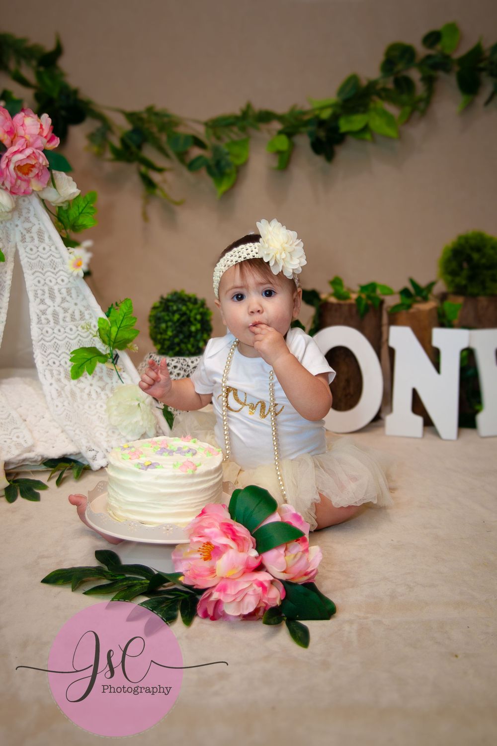 First birthday celebration setup with white cake, pink flowers, and greenery garland against neutral backdrop.