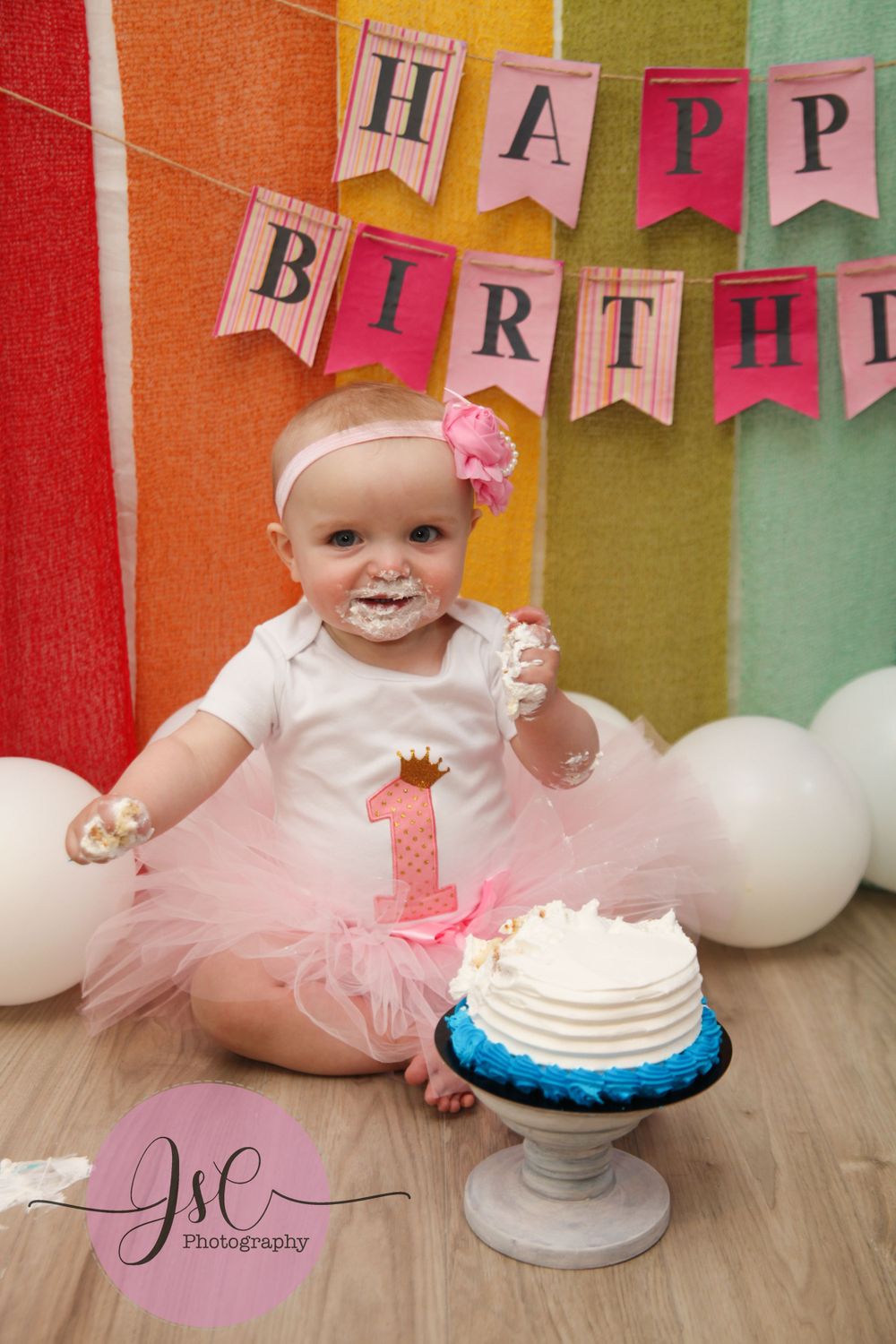 Colorful birthday banner hangs above baby enjoying first birthday cake smash session.