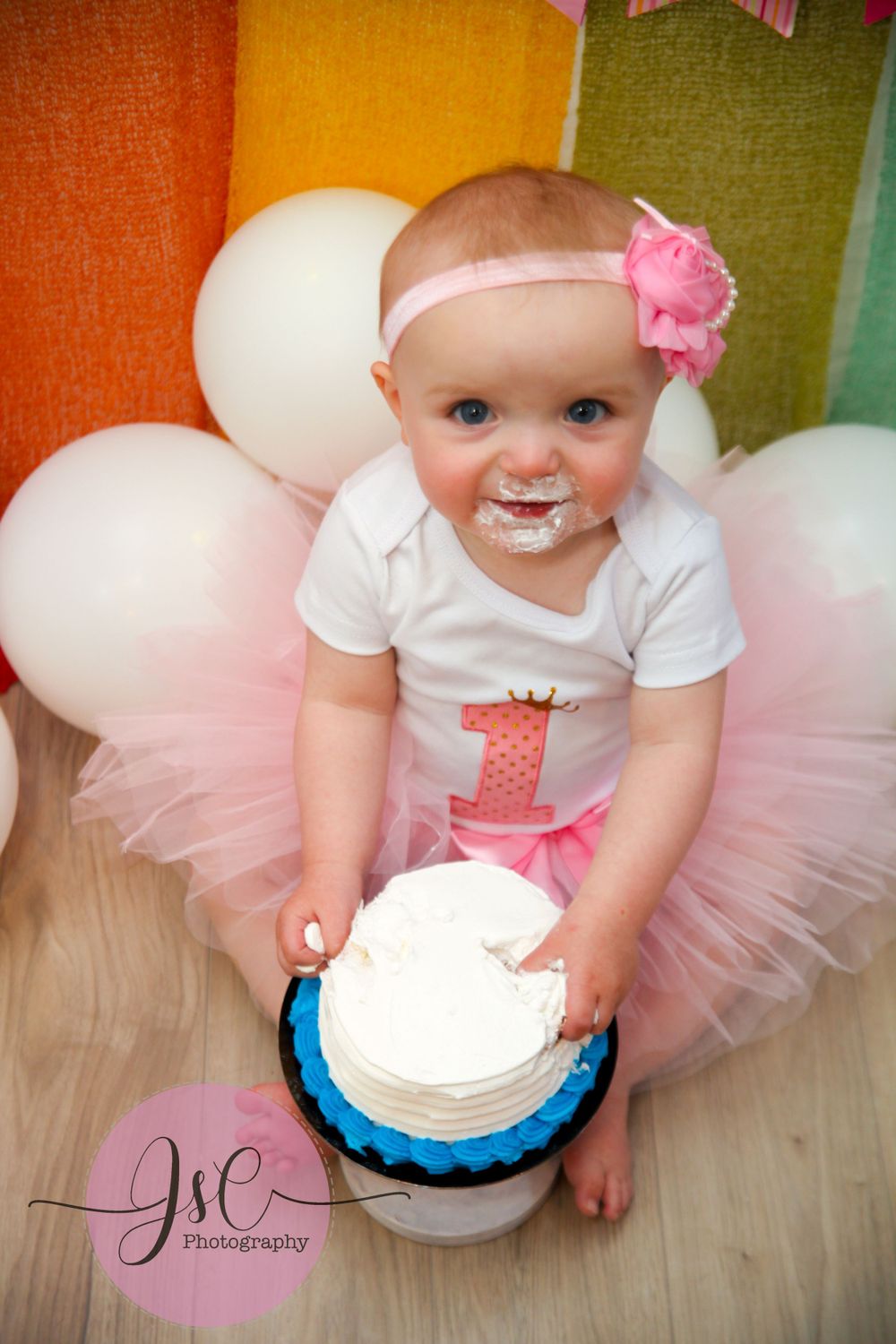 Baby in pink tutu dress reaching for white birthday cake on wooden floor with balloons.