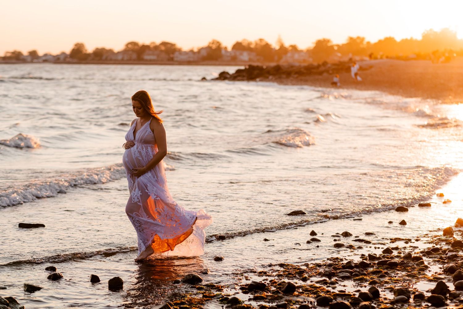 An expectant mother in a flowing dress walks along a beach at golden sunset.
