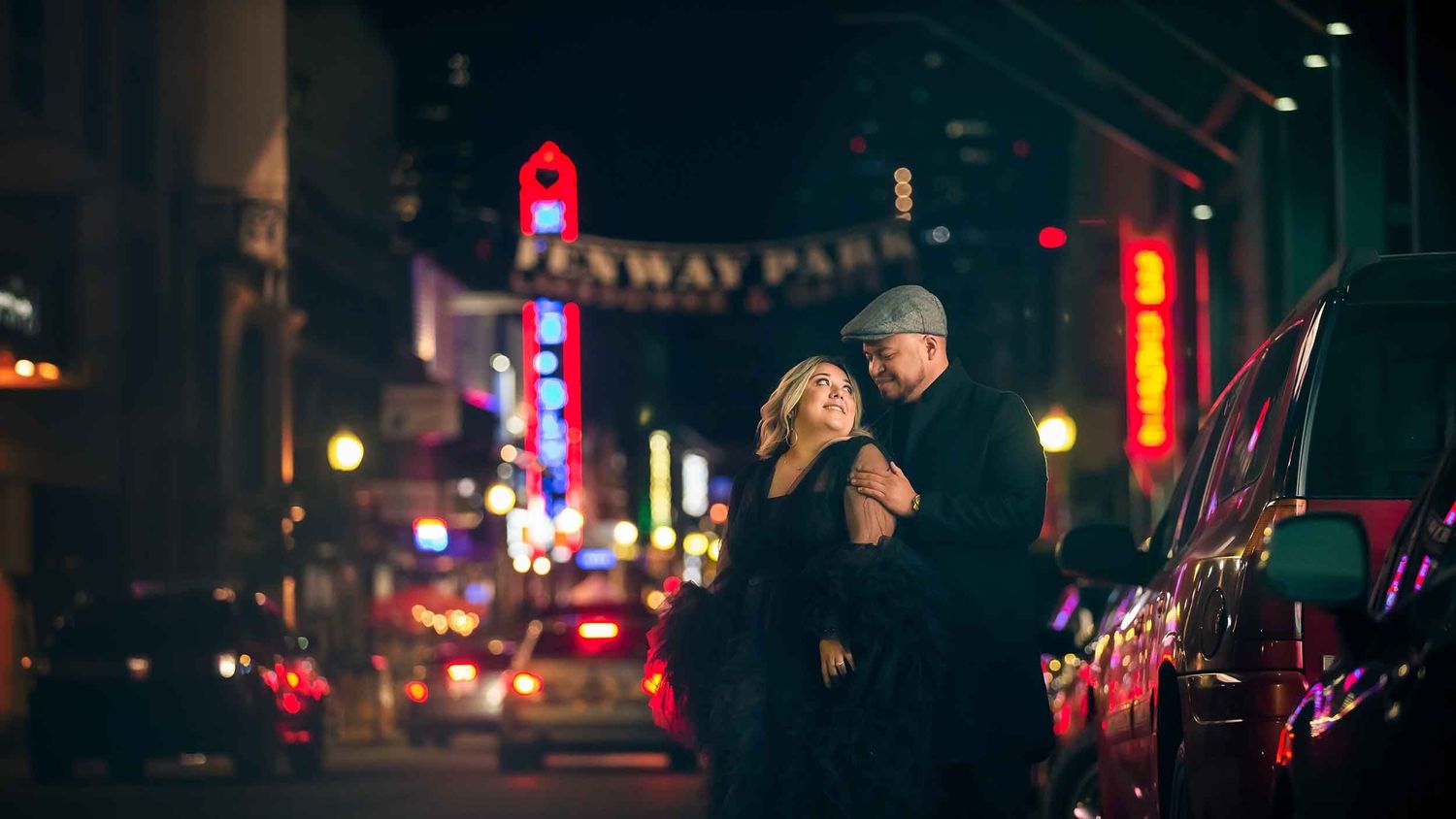Couple sharing a romantic nighttime engagement portrait in front of Fenway Park with glowing neon lights and a cinematic city atmosphere
