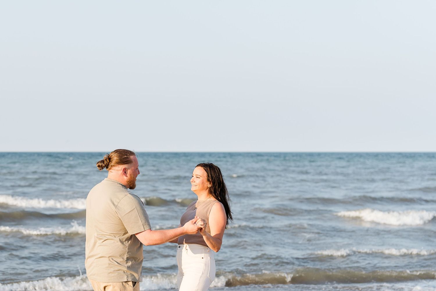 Couple holding hands and facing each other on a sunny beach with calm ocean waves in the background.