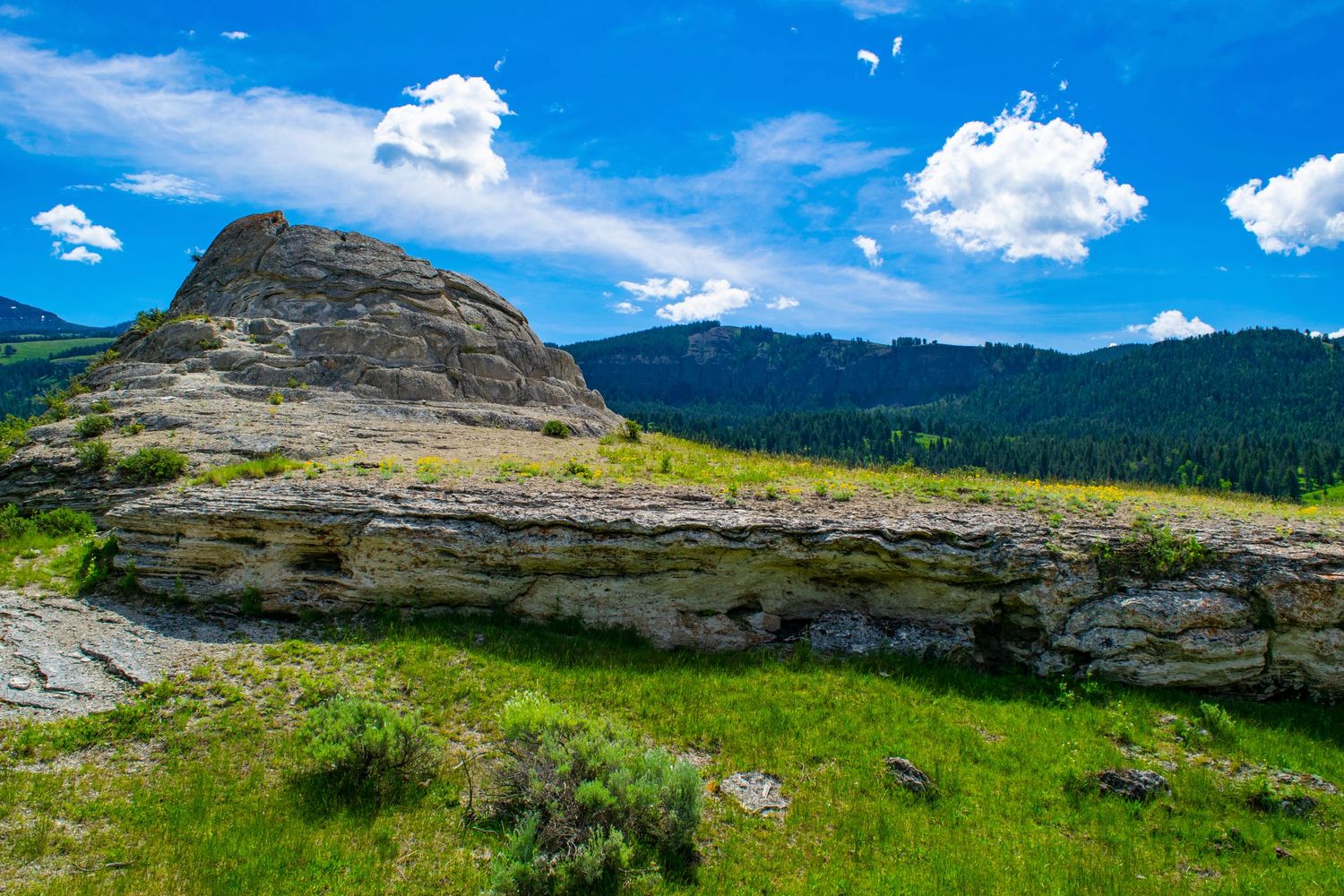 Large layered rock outcropping stands prominently against mountain backdrop and blue sky.