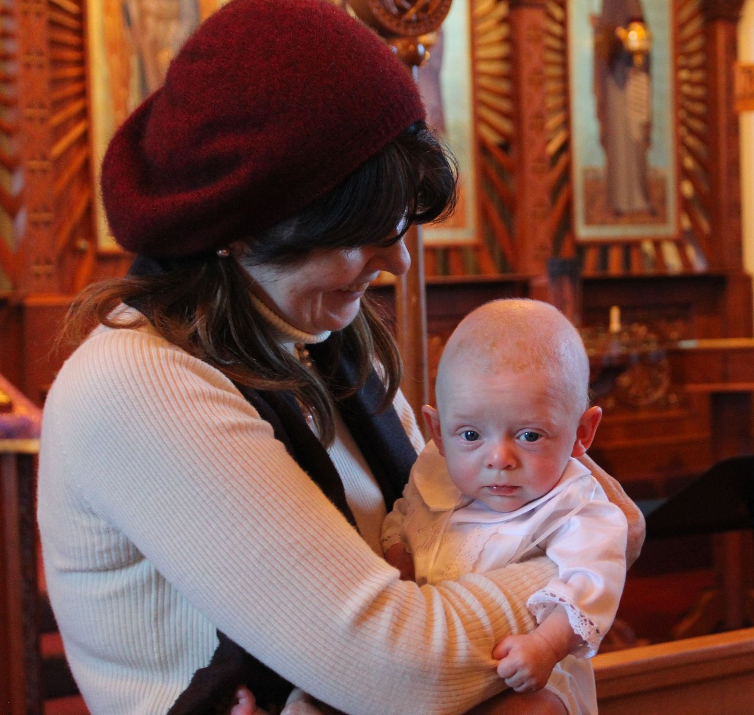 A baby in white attending a baptismal ceremony with religious icons in background.
