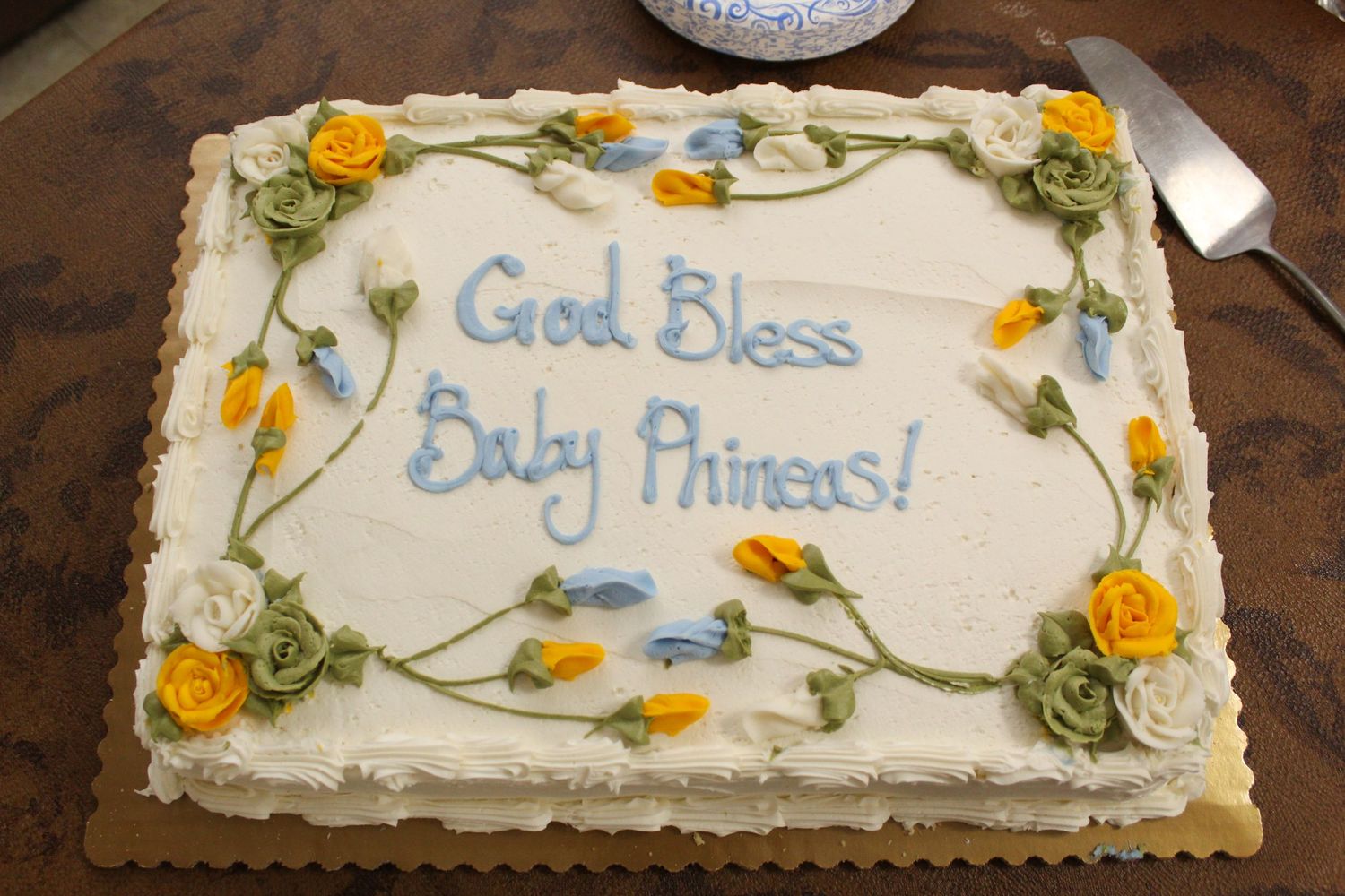 A decorated white cake with yellow flowers reading 'God Bless Baby Phineas'.