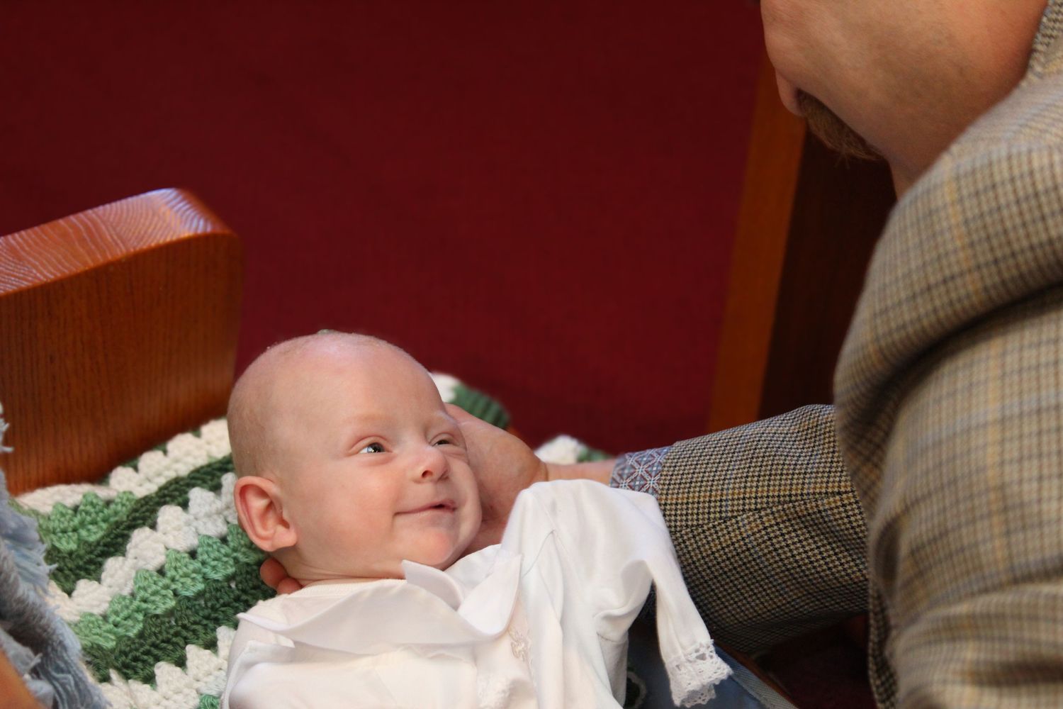 A smiling infant in white baptismal gown lying on a crocheted blanket.