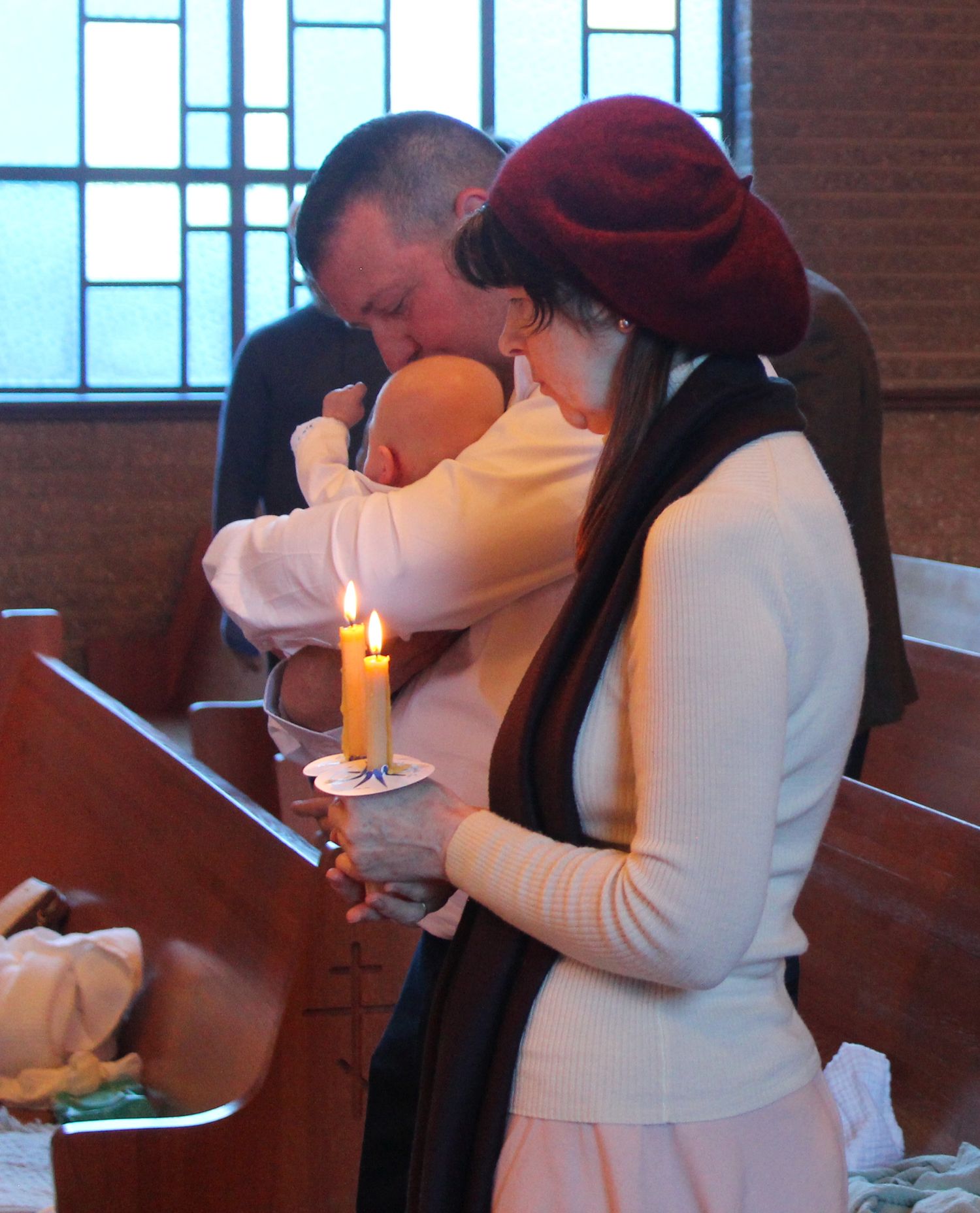 A baptism ceremony with a baby in white at a church altar with lit candles.