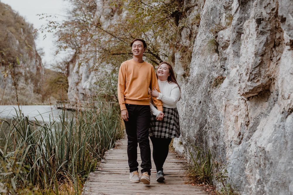 Wide shot of her holding his hand while they walk through Plitvice National Park, capturing their connection.