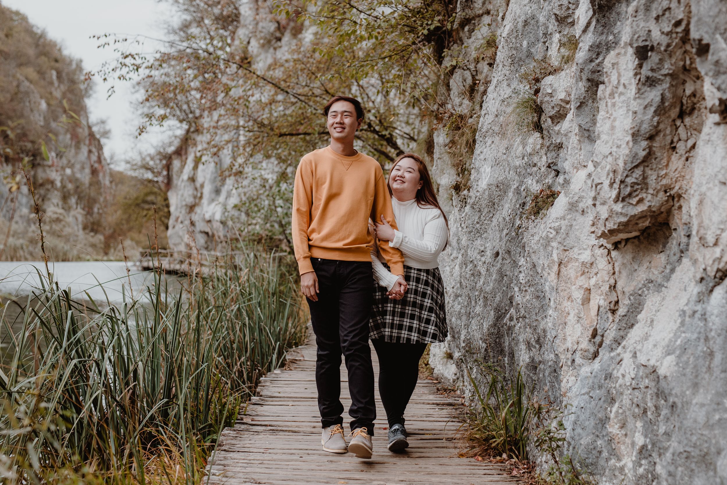 Wide shot of her holding his hand while they walk through Plitvice National Park, capturing their connection.