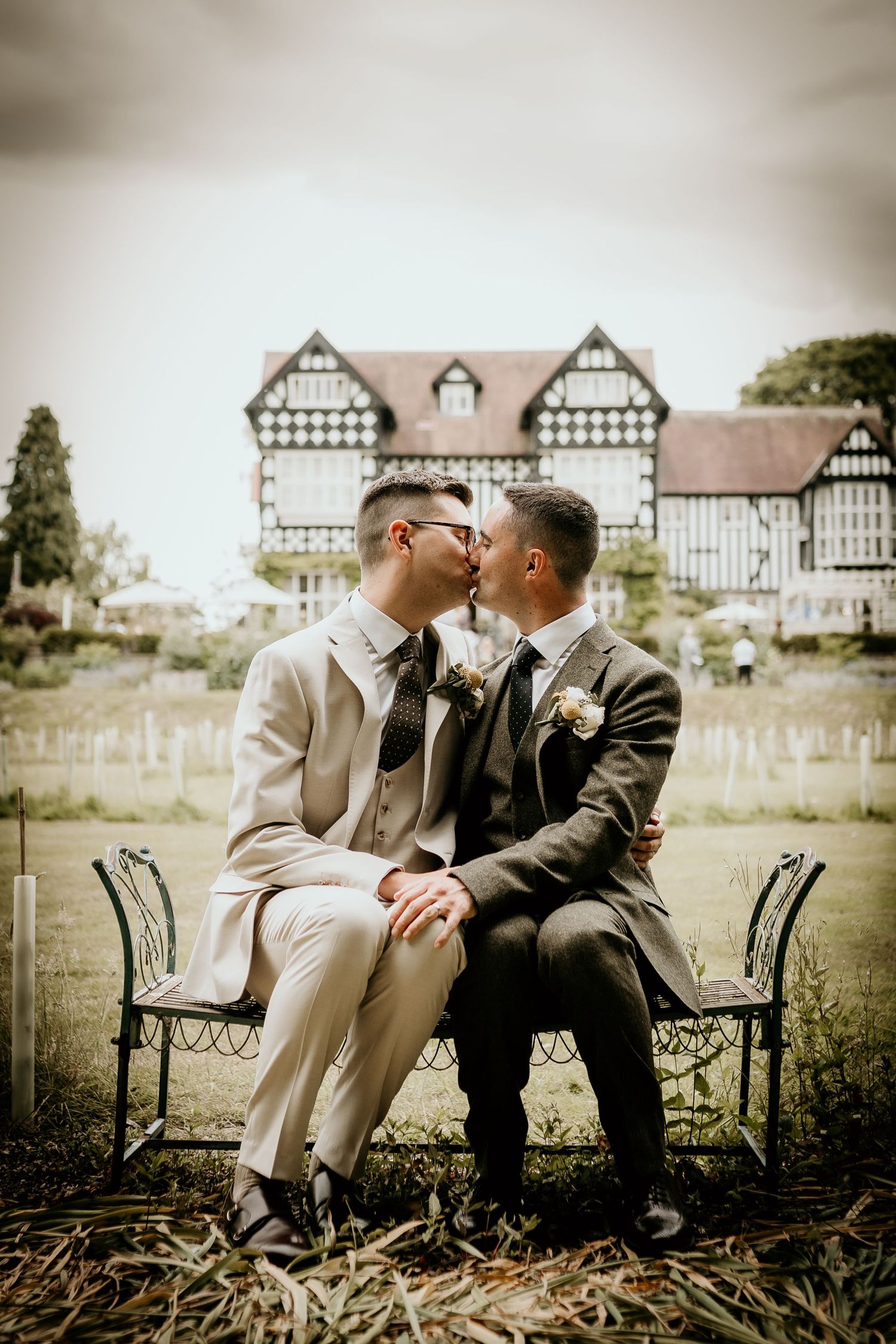 Two people share an intimate moment while seated on a garden bench with Tudor-style buildings in the background.