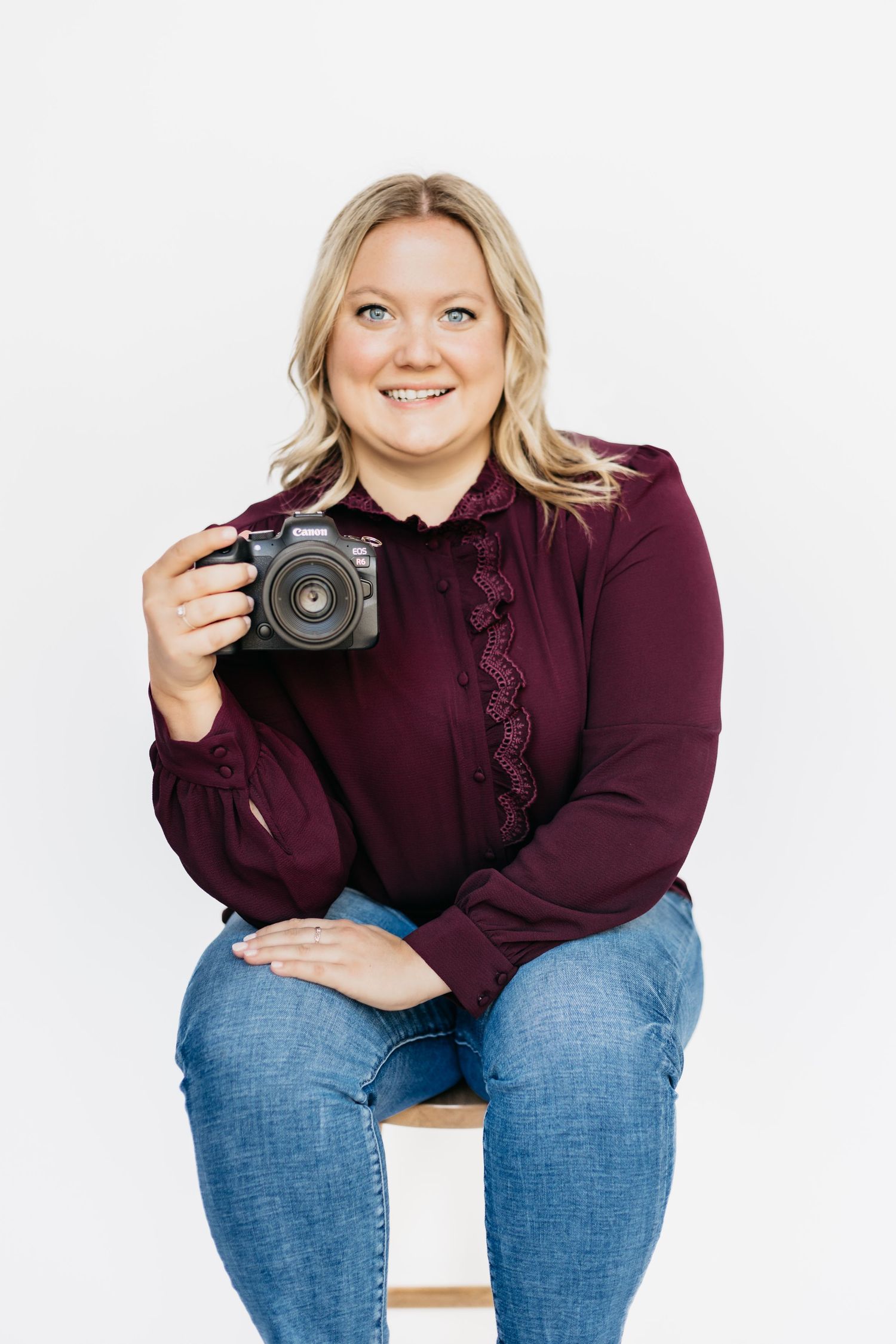 Your Photographer: Smiling blonde woman in burgundy blouse sitting on stool holding a mirrorless camera.