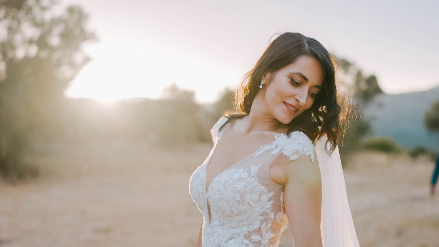 A bride in a lace wedding dress poses in golden sunlight against a desert landscape.