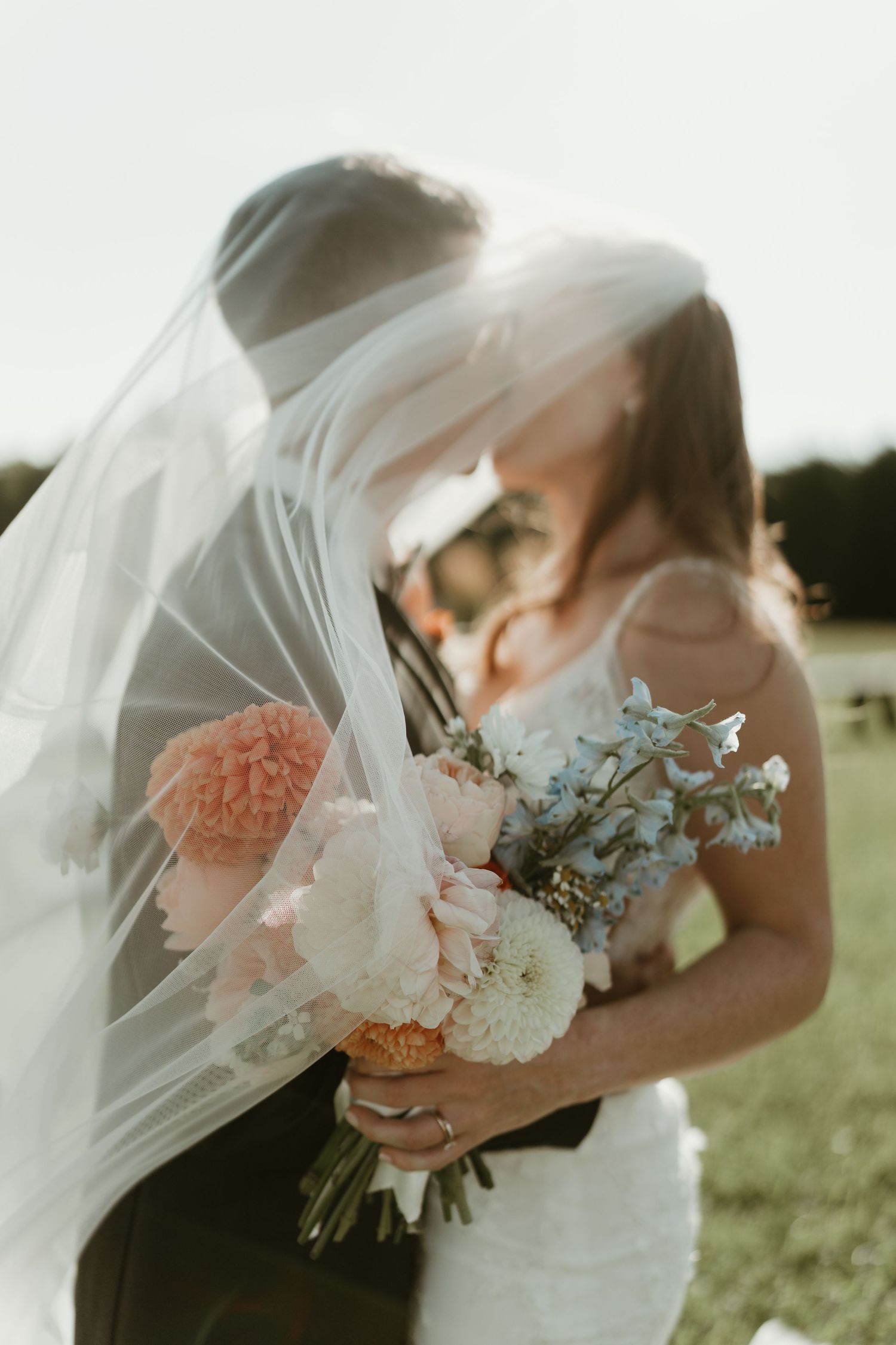 Newlyweds share an intimate moment behind a bridal bouquet with peach and white flowers.