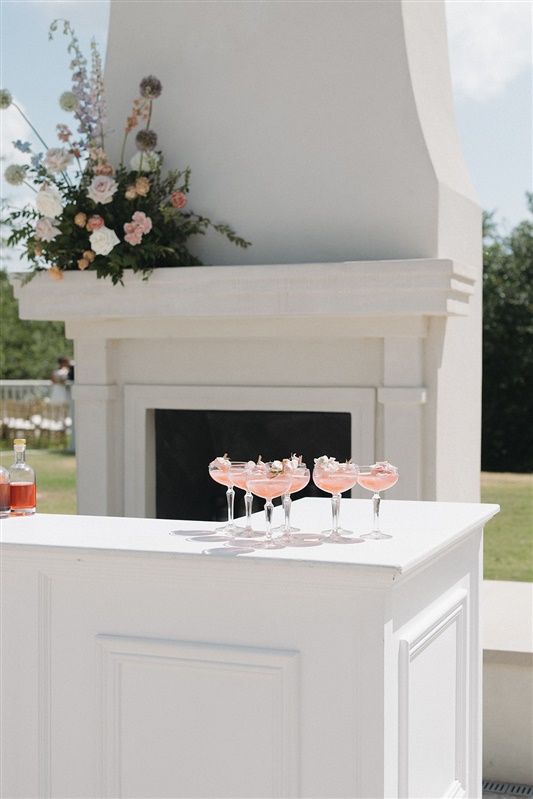 Pink cocktails in coupe glasses displayed on a white bar counter next to an elegant fireplace with floral arrangements.