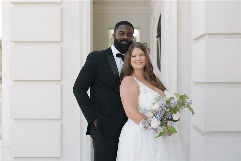 A bride and groom stand together in front of white pillars, the bride holding a delicate bouquet of purple and white flowers.