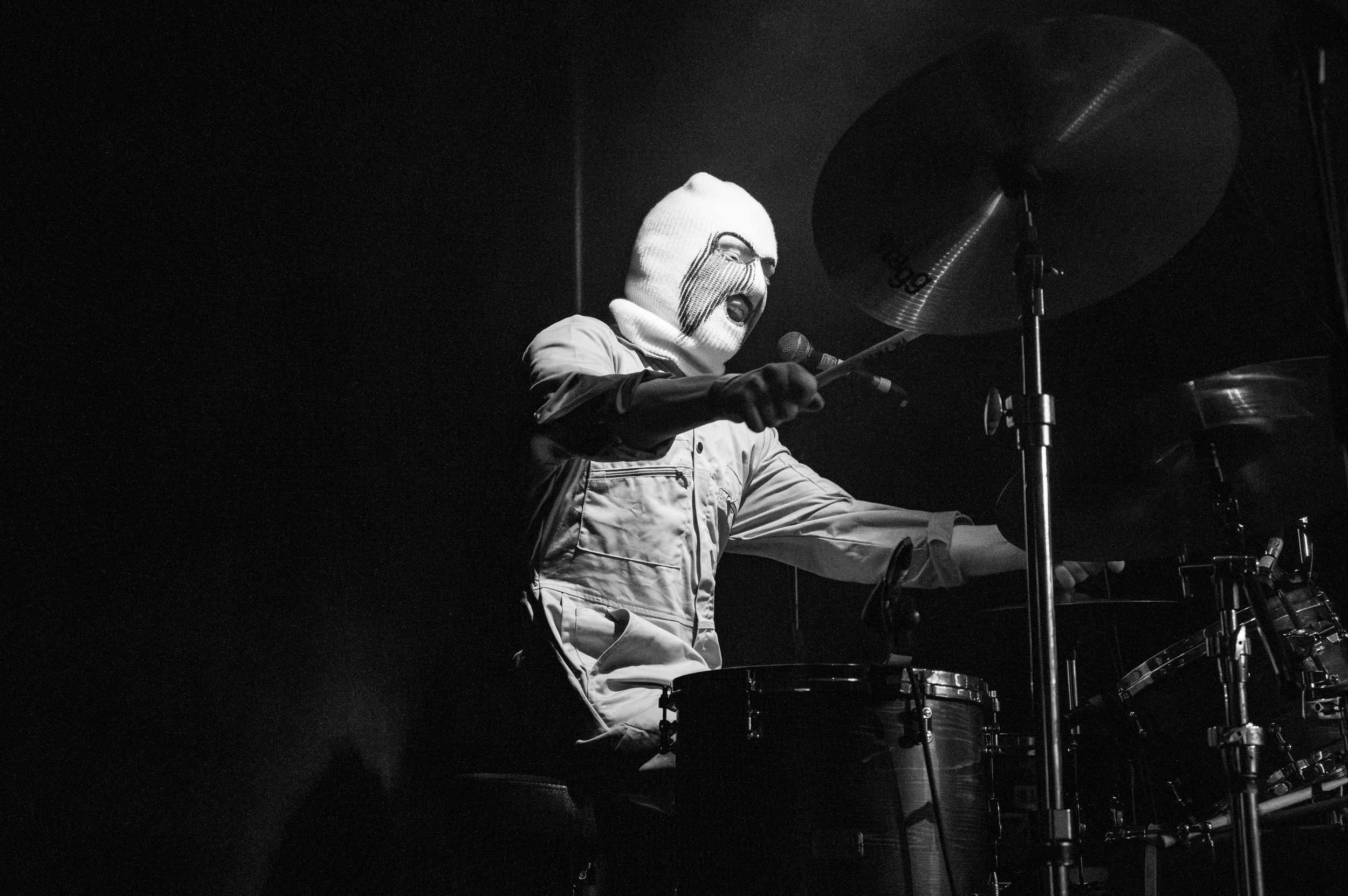 Drummer performs with intensity during live concert in dramatic black and white lighting.