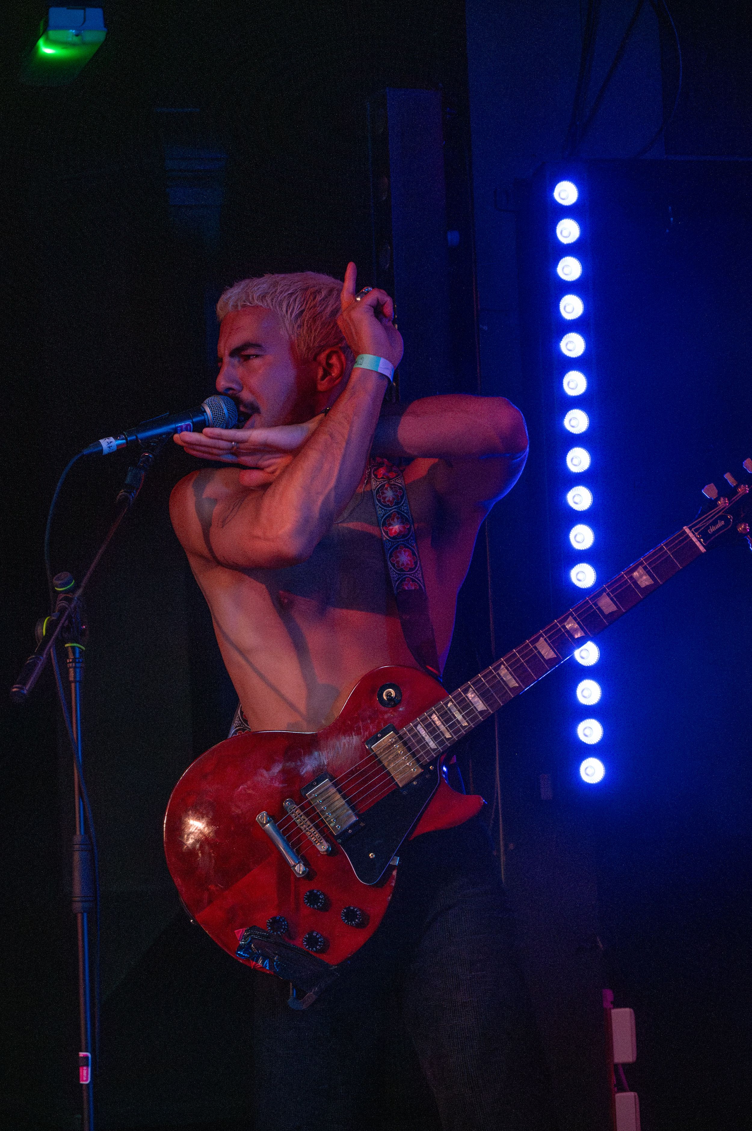Shirtless musician plays red guitar under blue stage lights during concert performance.