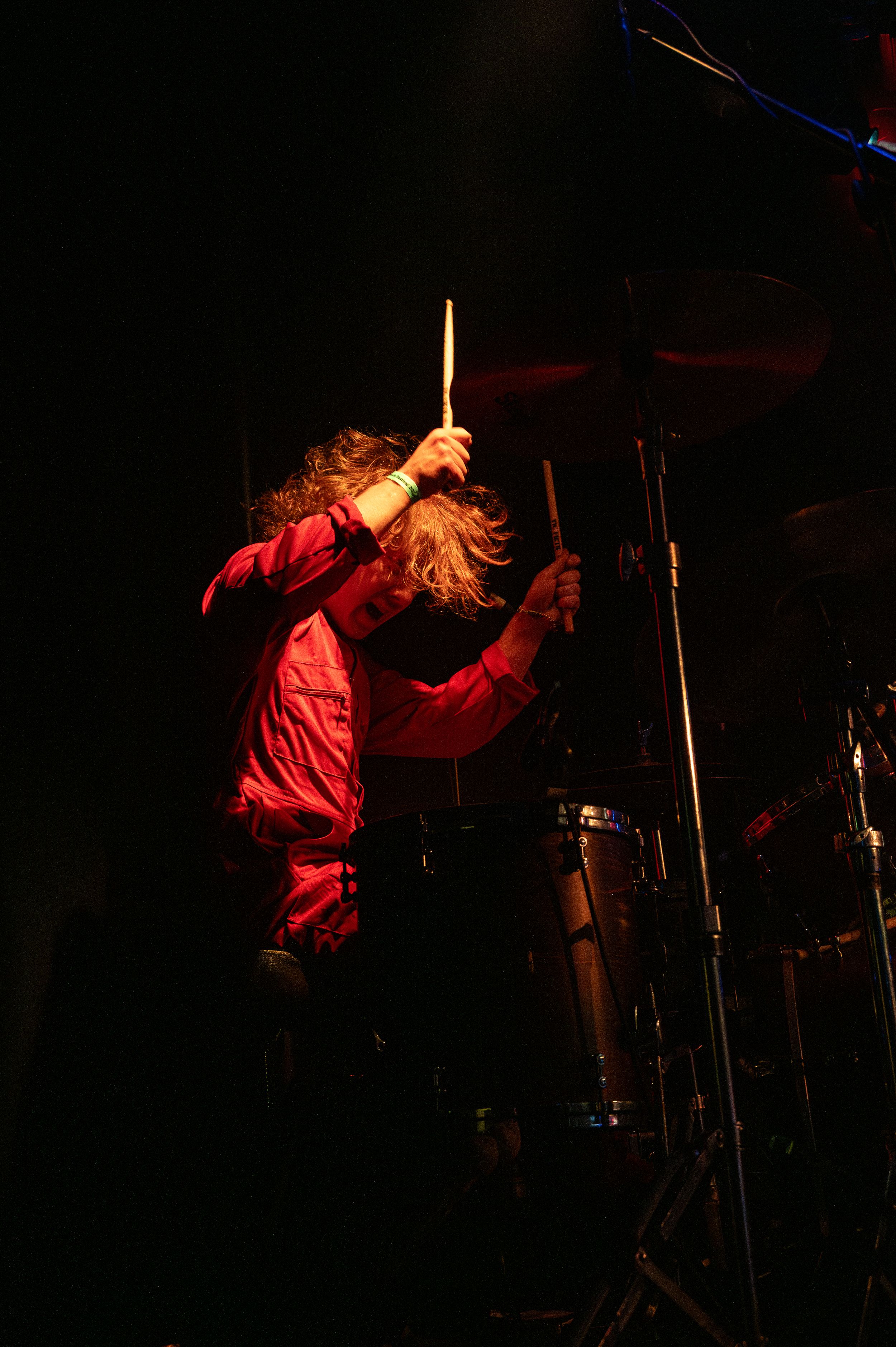 Performer in red clothing plays drums with hair flying during dramatic low-light concert moment.