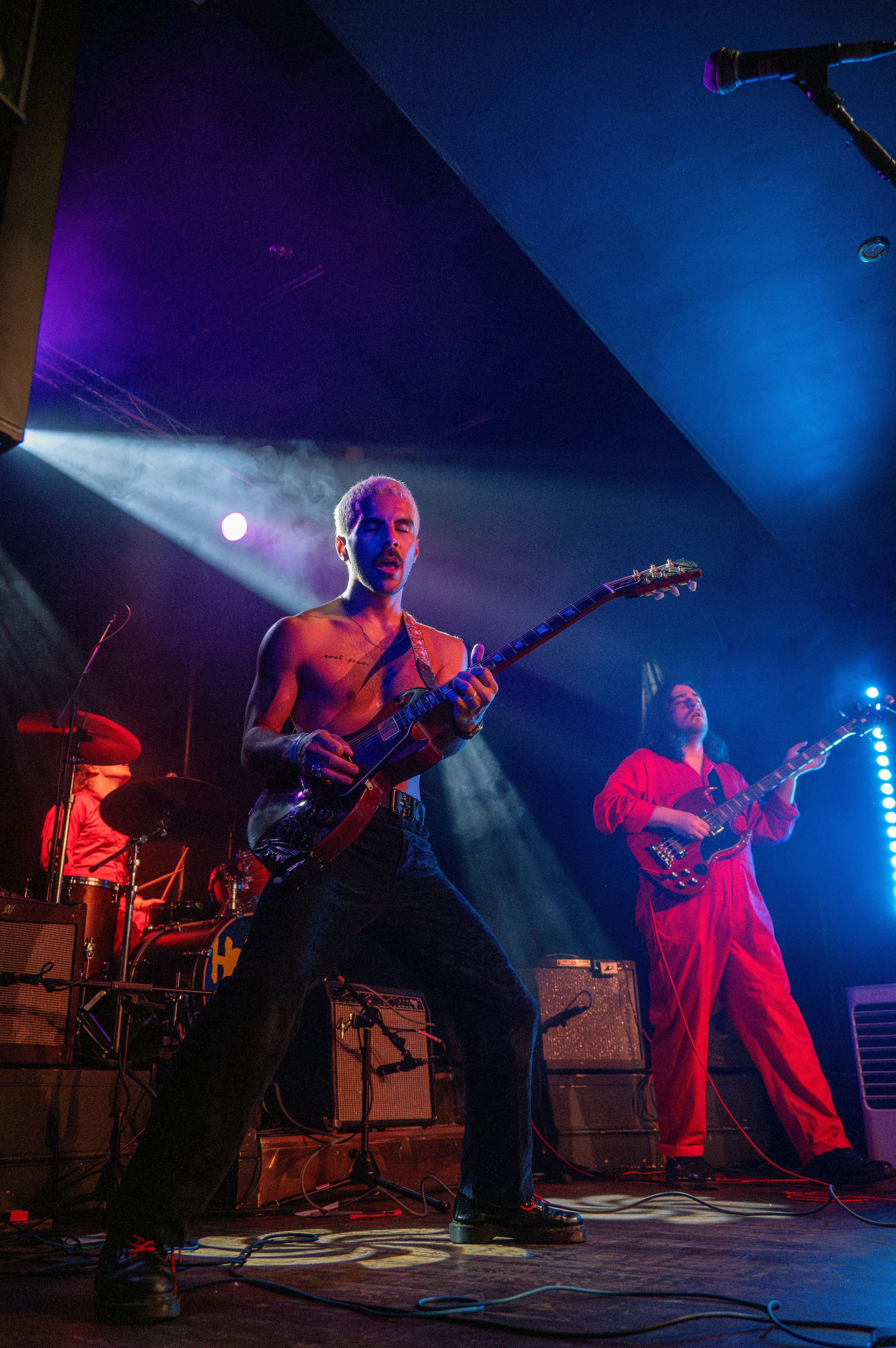 Shirtless performers playing guitars on stage under vibrant blue and purple lighting.