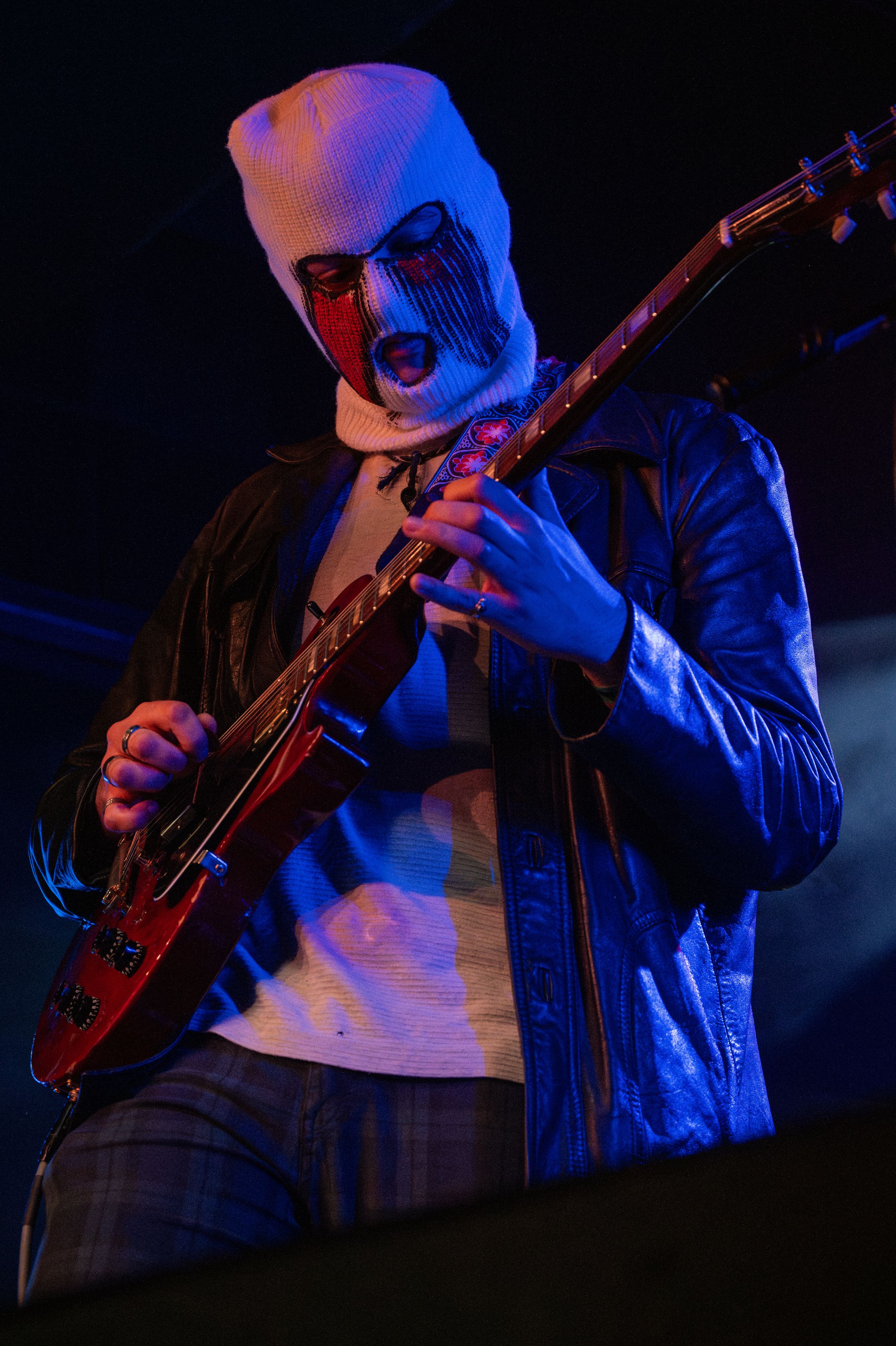 Guitarist wearing leather jacket and white mask performing under dramatic stage lighting.