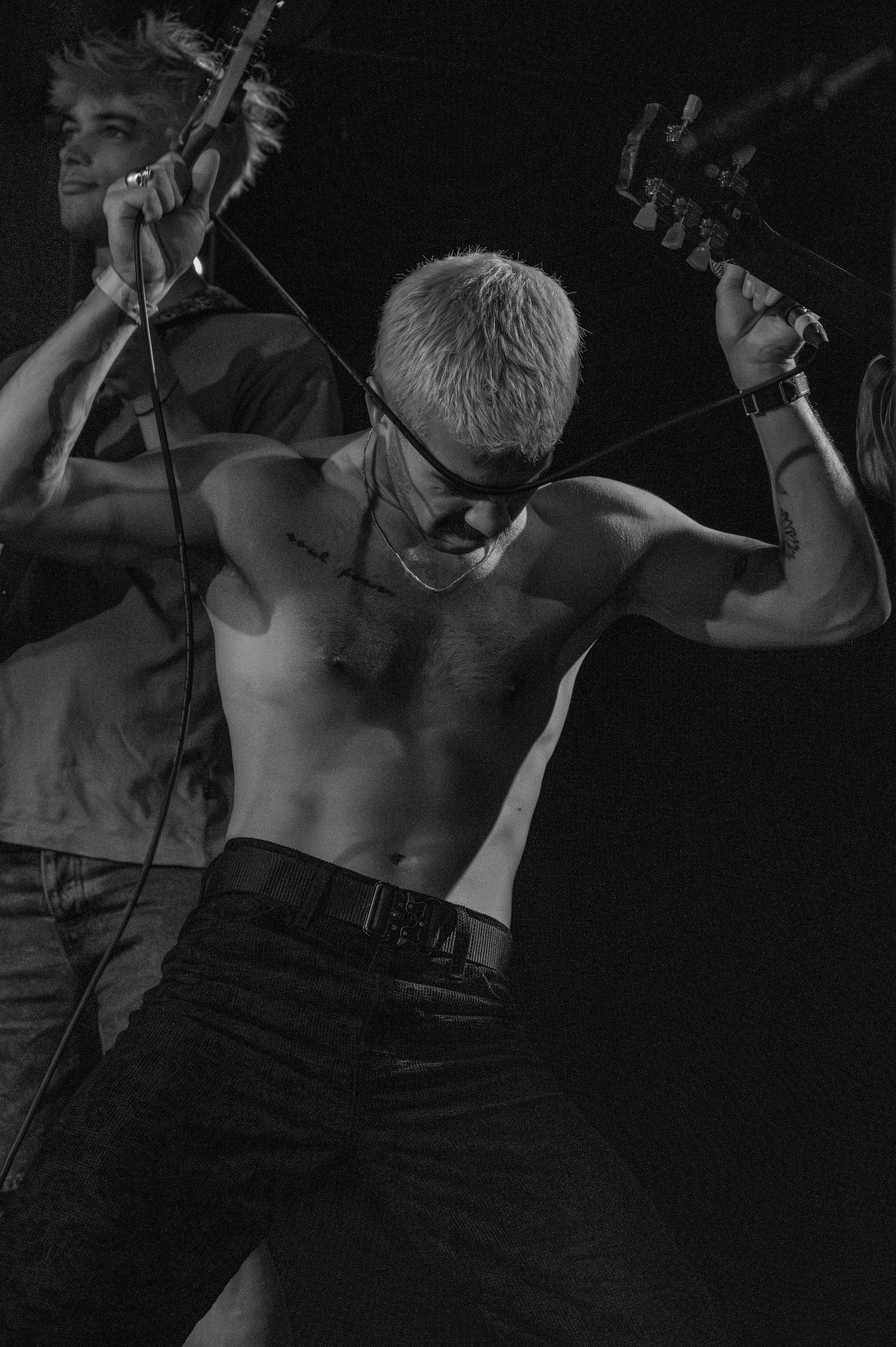 Black and white photo of shirtless performer playing guitar on dark stage.