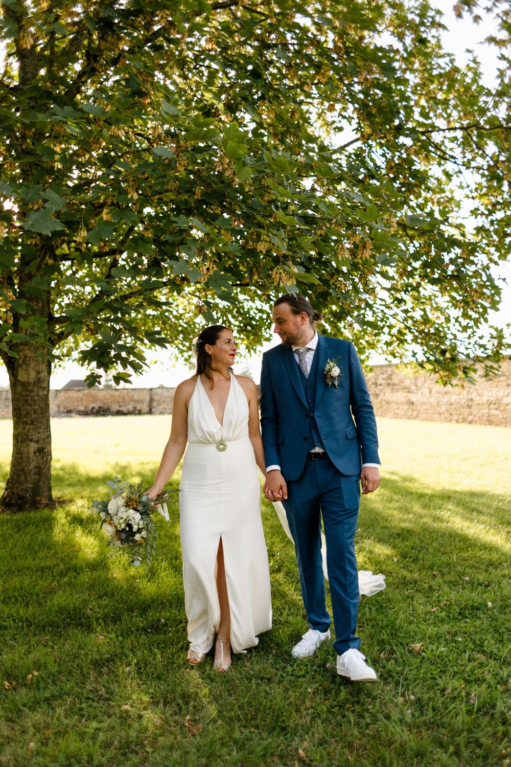 A couple in wedding attire walks hand in hand through a lush green garden setting under sunlit trees.