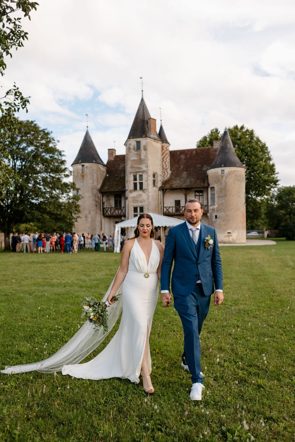 A couple walks on green grass with a historic French chateau with turrets and towers in the background.