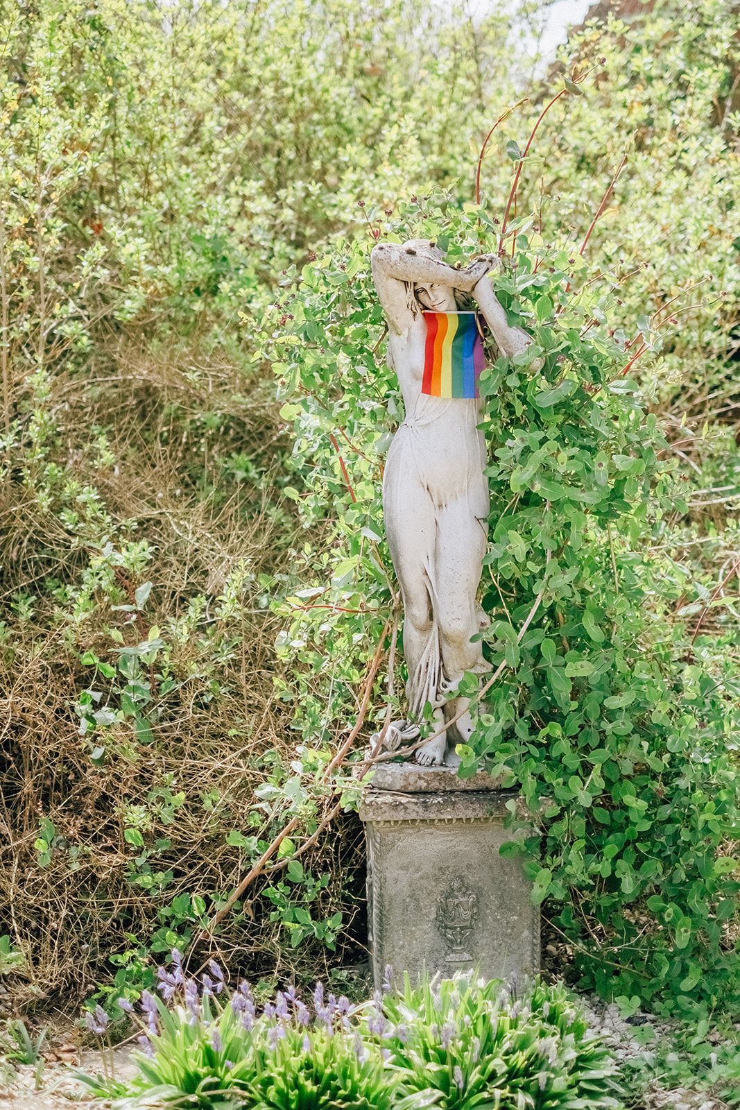 A classical garden statue with a rainbow scarf draped around its neck stands on a stone pedestal surrounded by greenery.