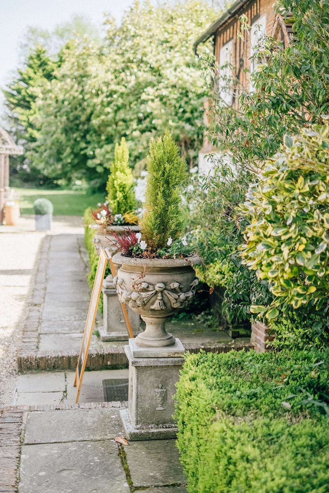 Ornate garden path with stone planters and decorative urns lined with lush greenery and trimmed hedges.