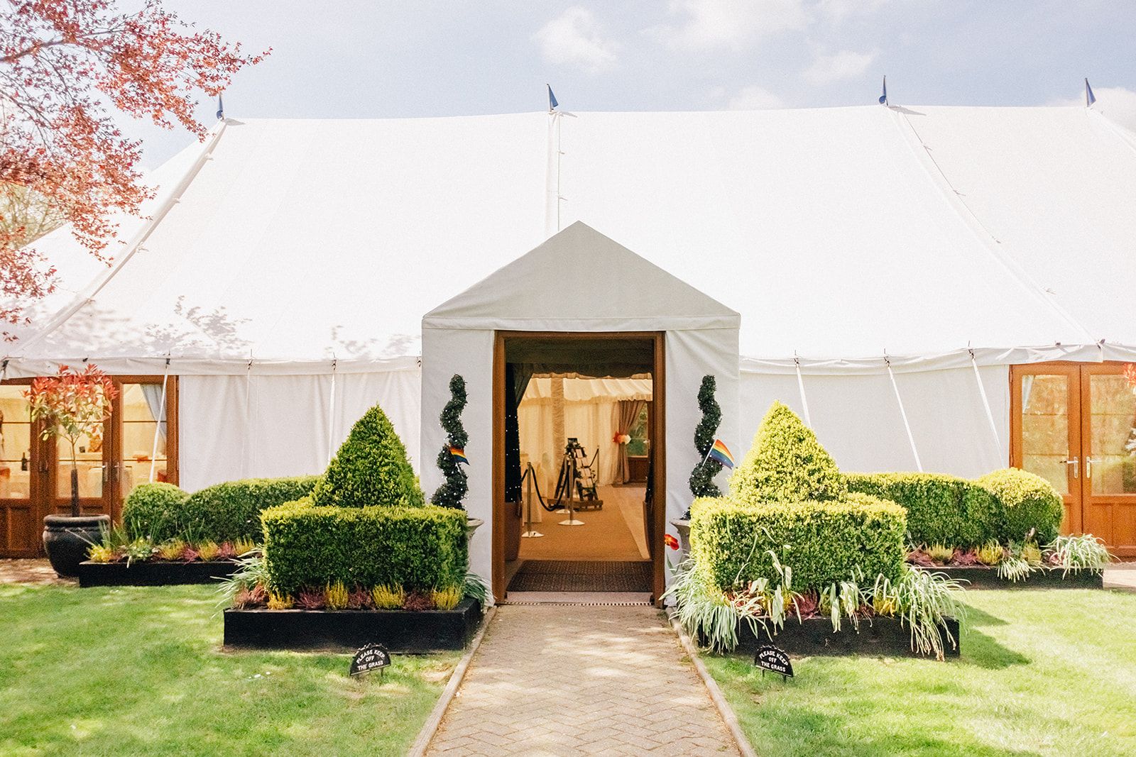 Elegant white wedding tent entrance with topiary bushes and formal garden landscaping.