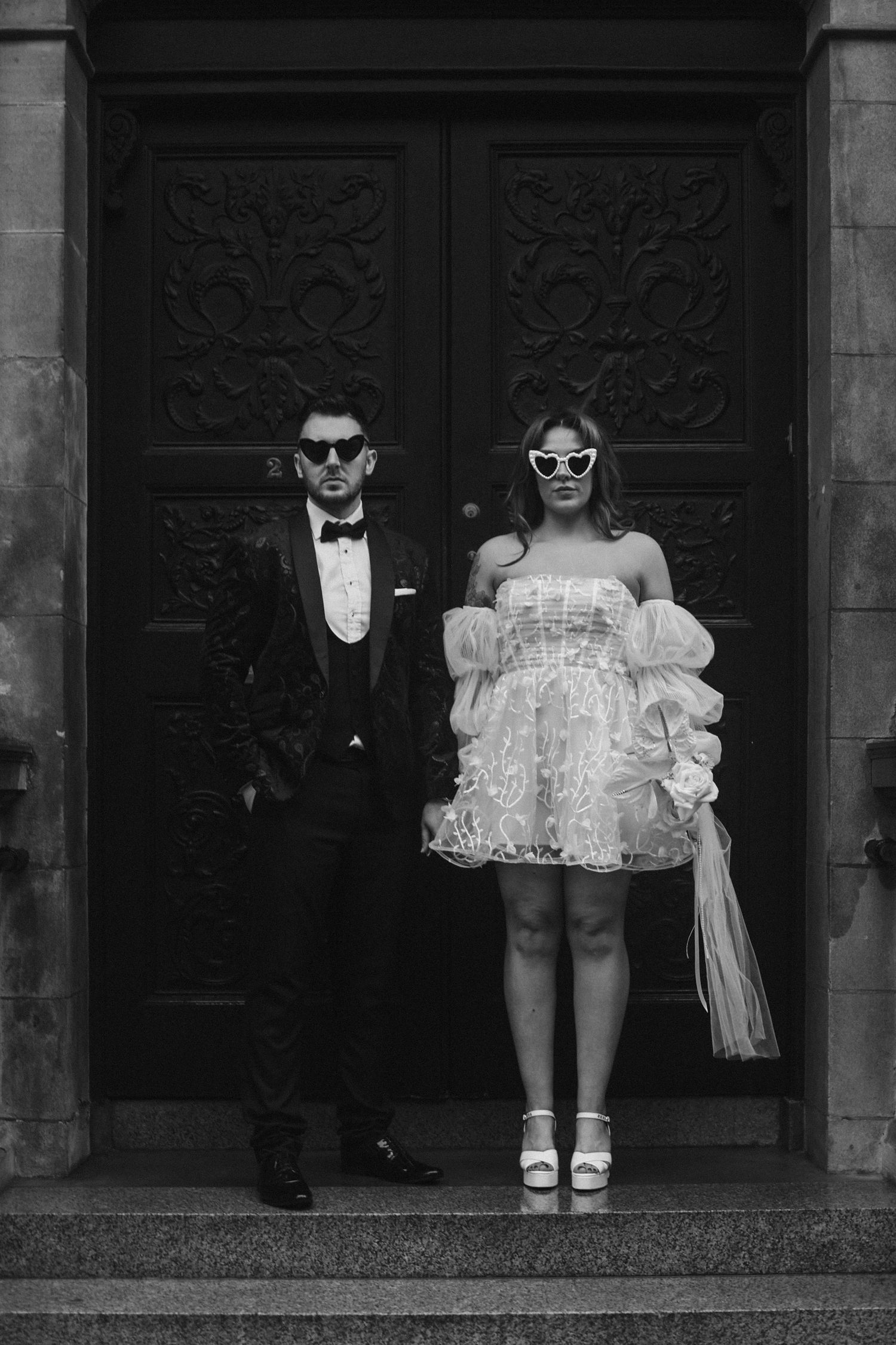 A stylish couple in sunglasses pose in front of ornate dark doors.