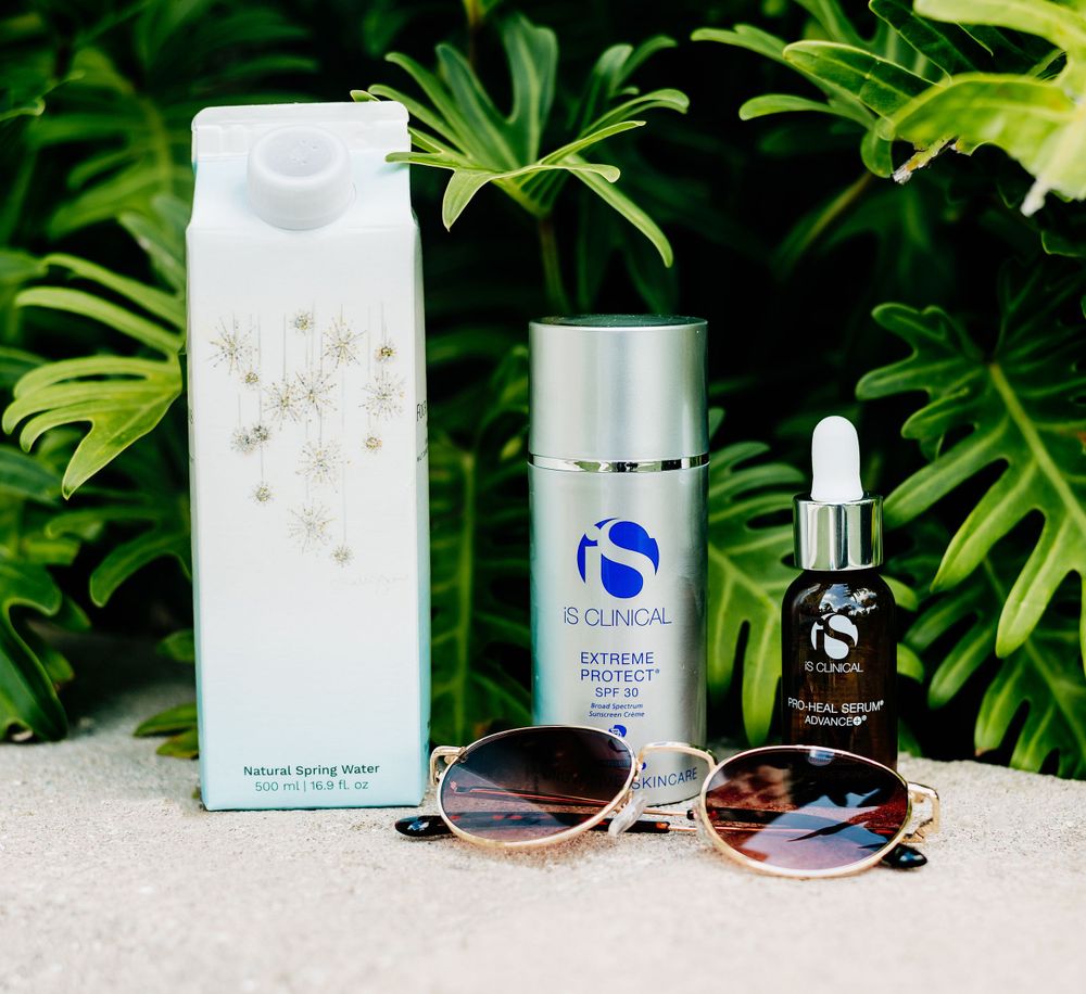 Beauty and skincare products arranged with sunglasses against a tropical plant background.