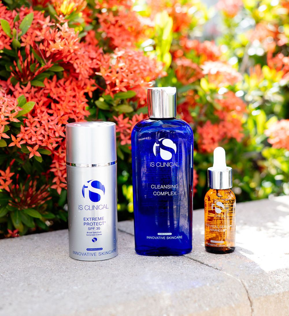 Three iS Clinical skincare products displayed against vibrant coral flowers in an outdoor setting.