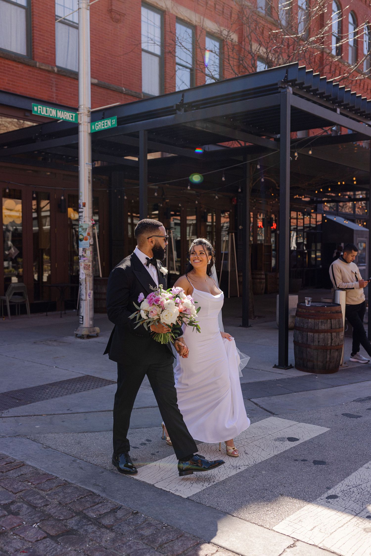Couple in wedding attire posing on urban street corner with brick building background.