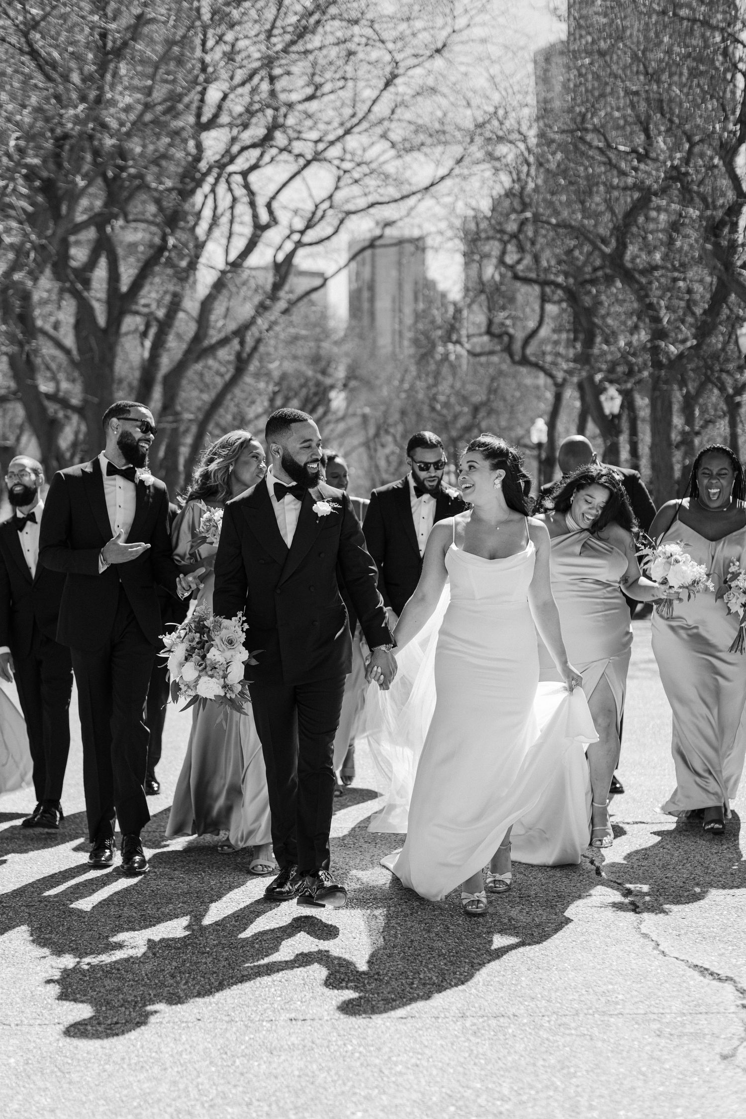 Wedding party walks through park in black and white with city buildings visible through trees.