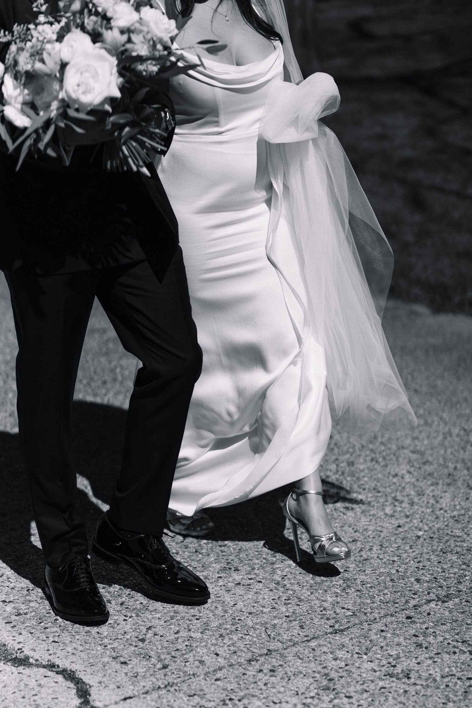 Black and white photo showing feet of couple walking, with bride in elegant white dress and flowing veil.