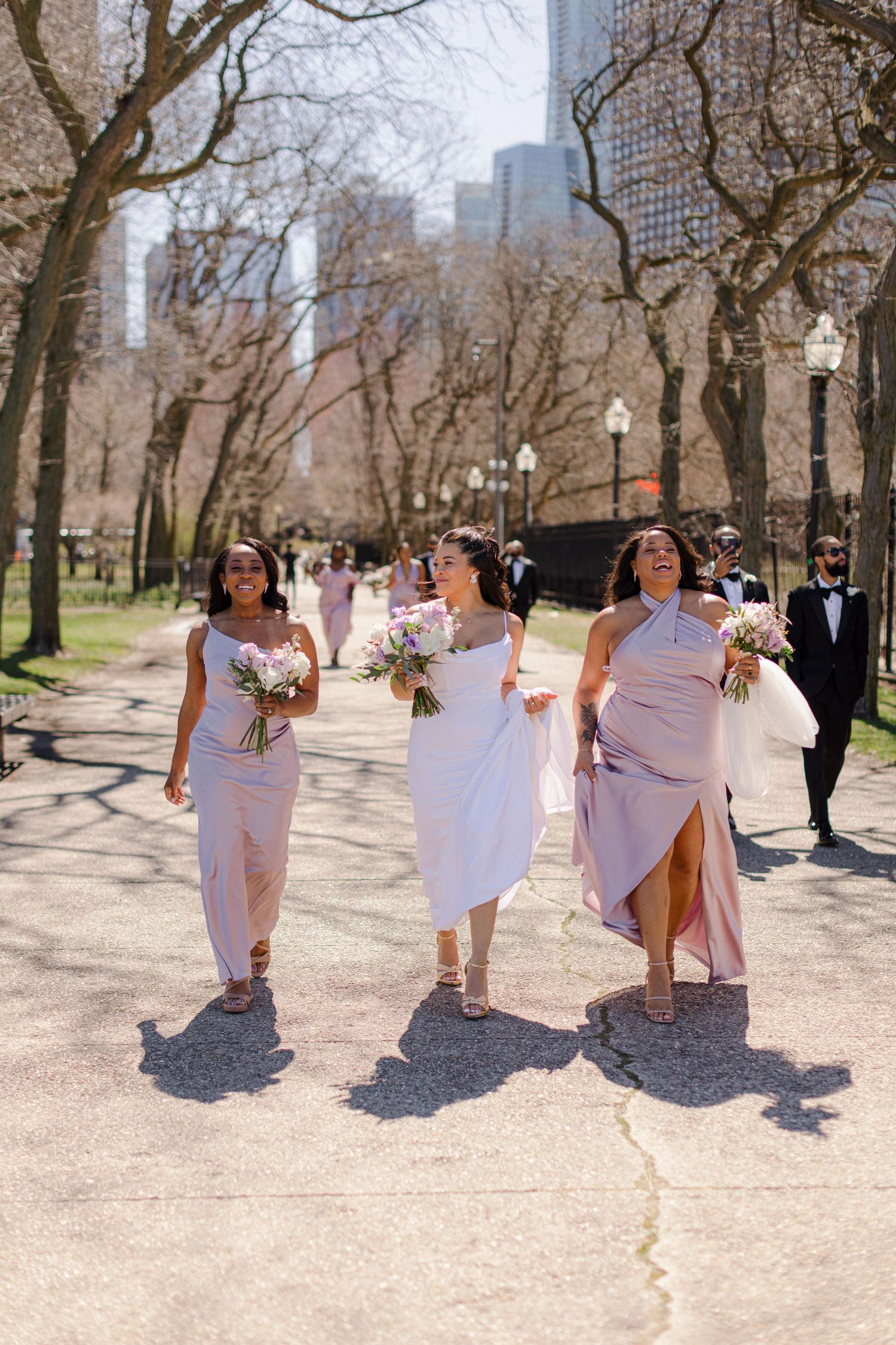 Bridesmaids in pink dresses walk through park with bouquets and city skyline in background.