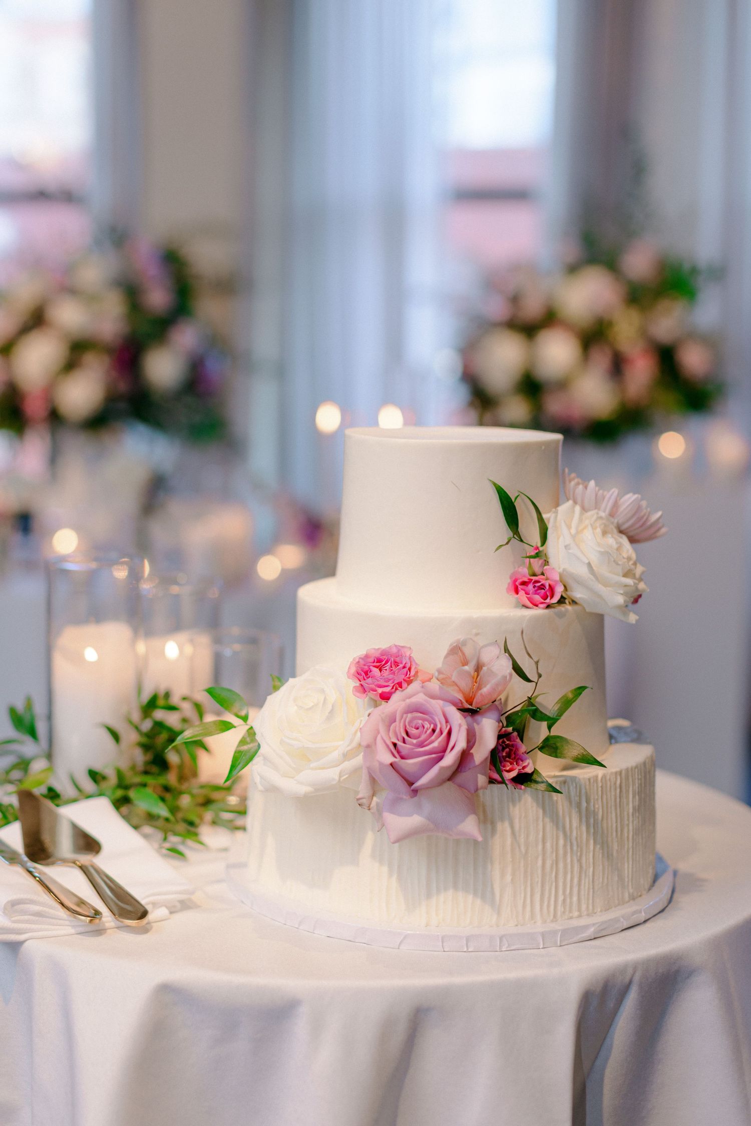 Elegant white wedding cake decorated with fresh pink and white roses on a white tablecloth.