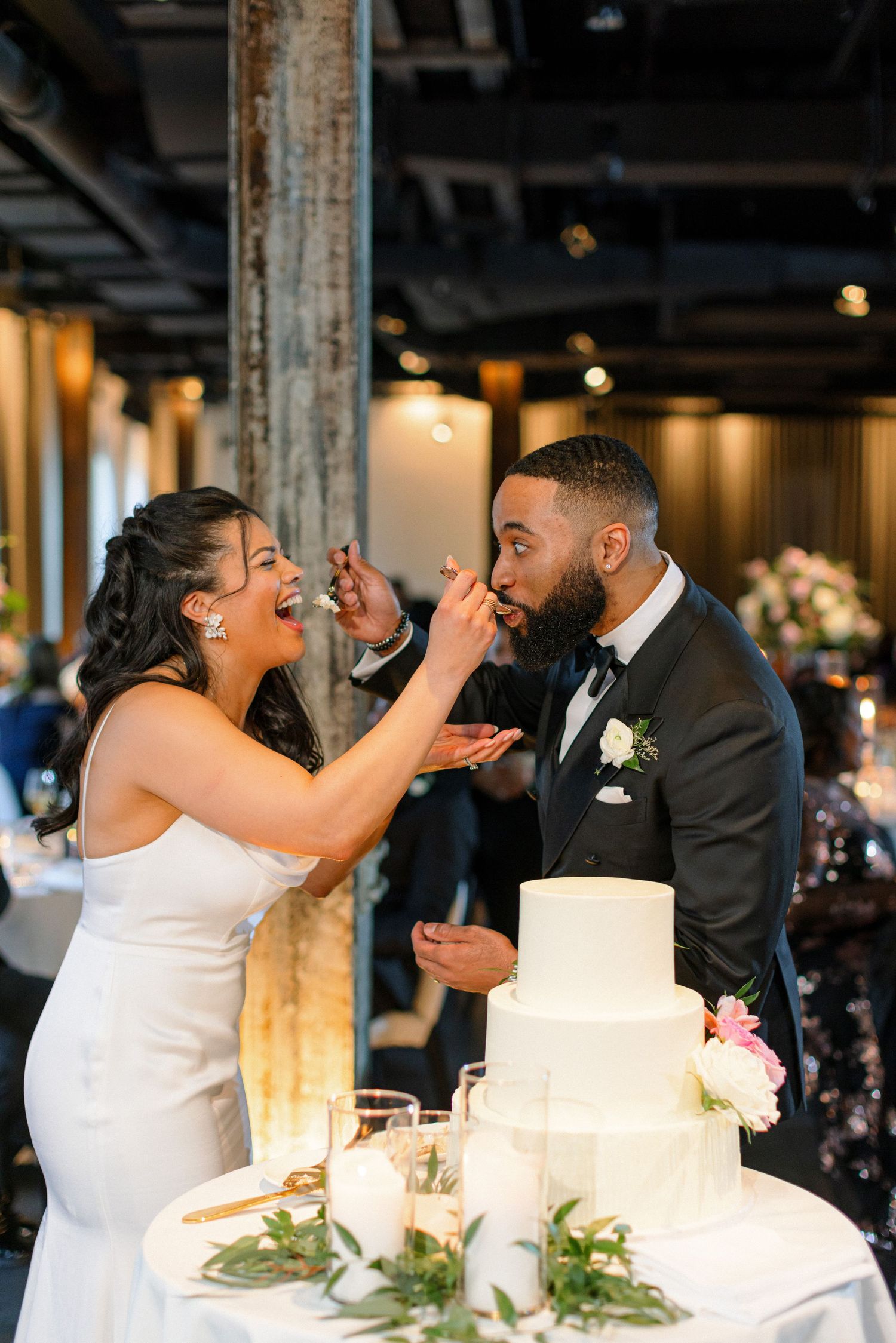 Playful newlyweds feeding each other wedding cake in a candid reception moment.