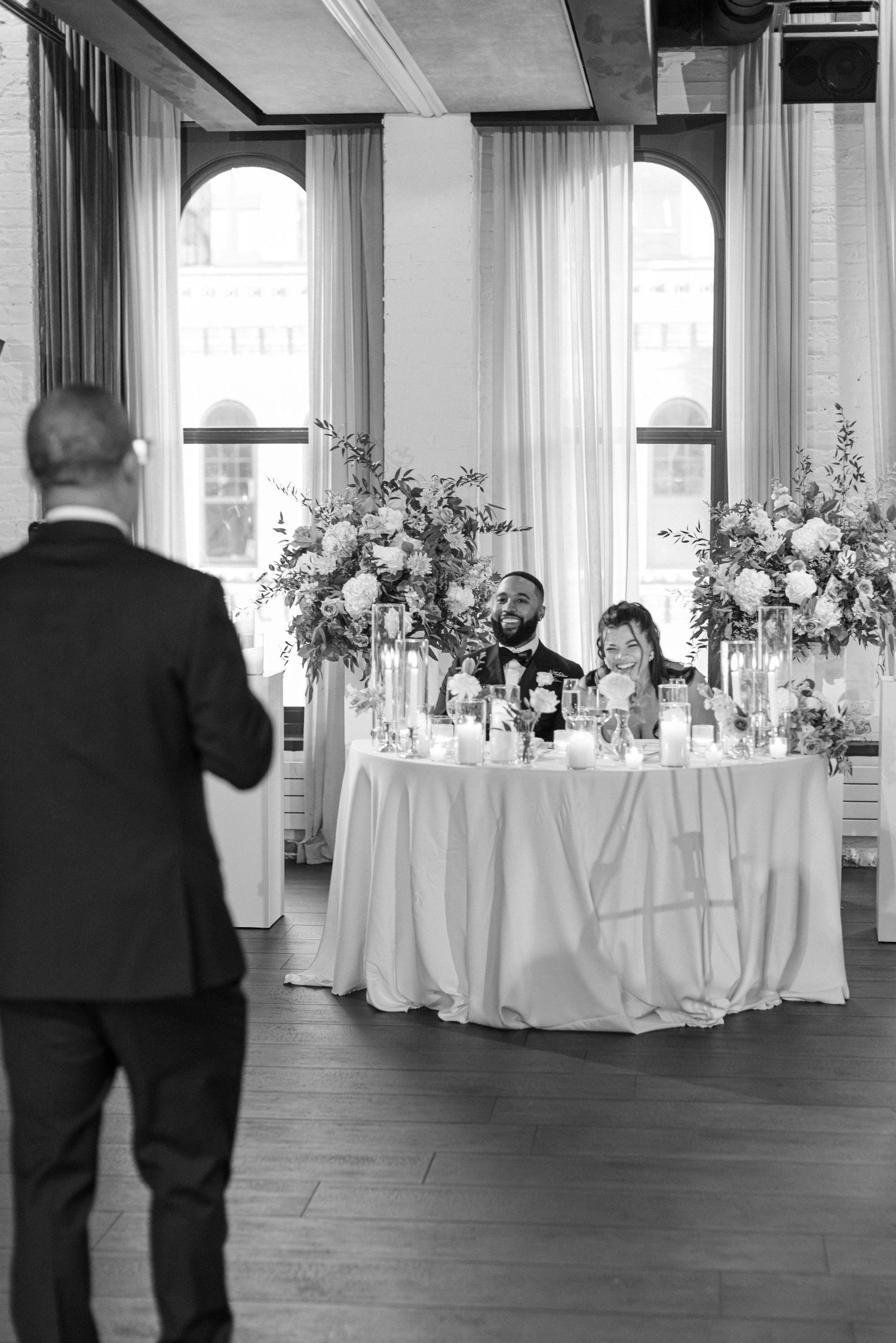 Black and white photo of wedding reception with elegant floral centerpieces and guests.