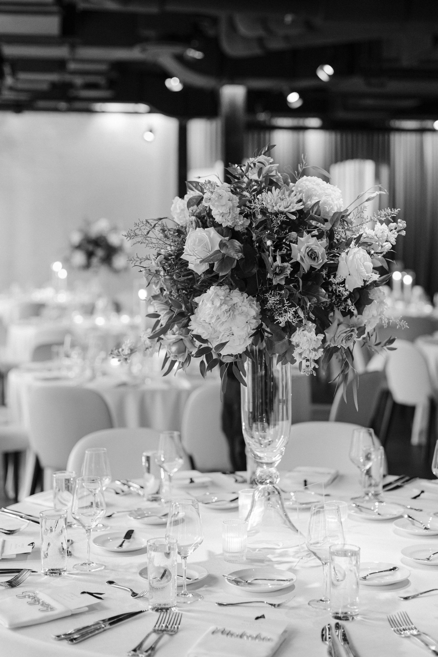 Black and white photo of an elegant wedding centerpiece with lush florals in a tall glass vase.