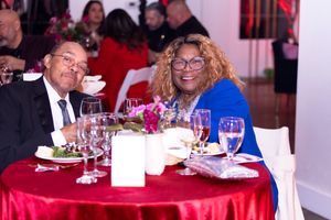 Guests enjoying dinner at round table with red tablecloth and floral centerpiece.