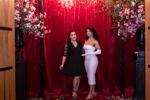 Two guests pose against red velvet backdrop with floral arrangements at formal event.