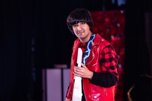 A performer in a red and black plaid jacket on stage with dramatic lighting.