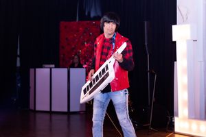 Musician in red jacket performs with a keyboard on stage with dramatic lighting.